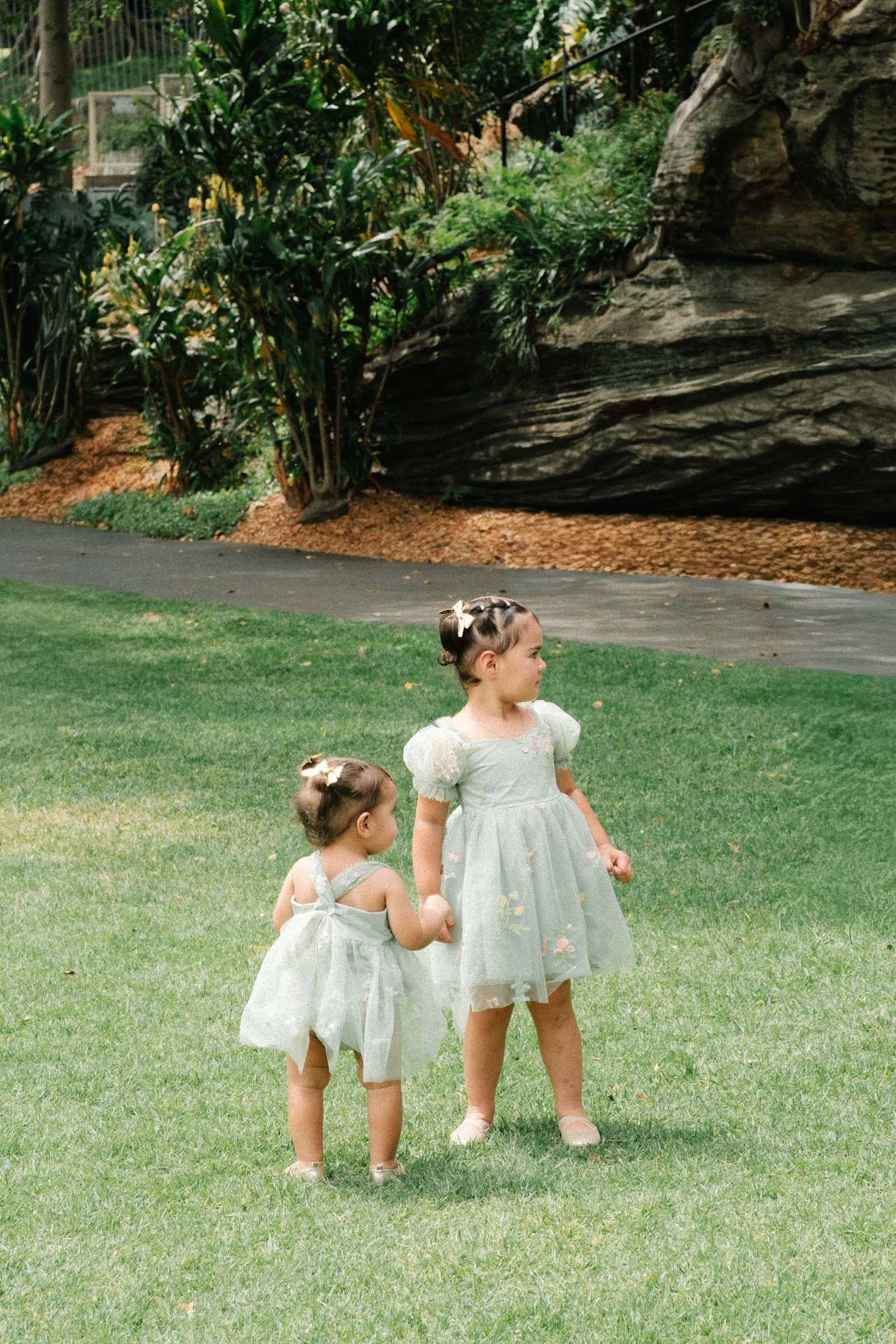 Two small children in white dresses crawl and explore together on a green lawn in a garden setting.