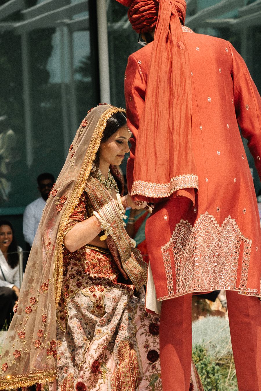 Traditional Indian bride in ornate gold and coral attire sits beside groom in matching formal wear during wedding ceremony.