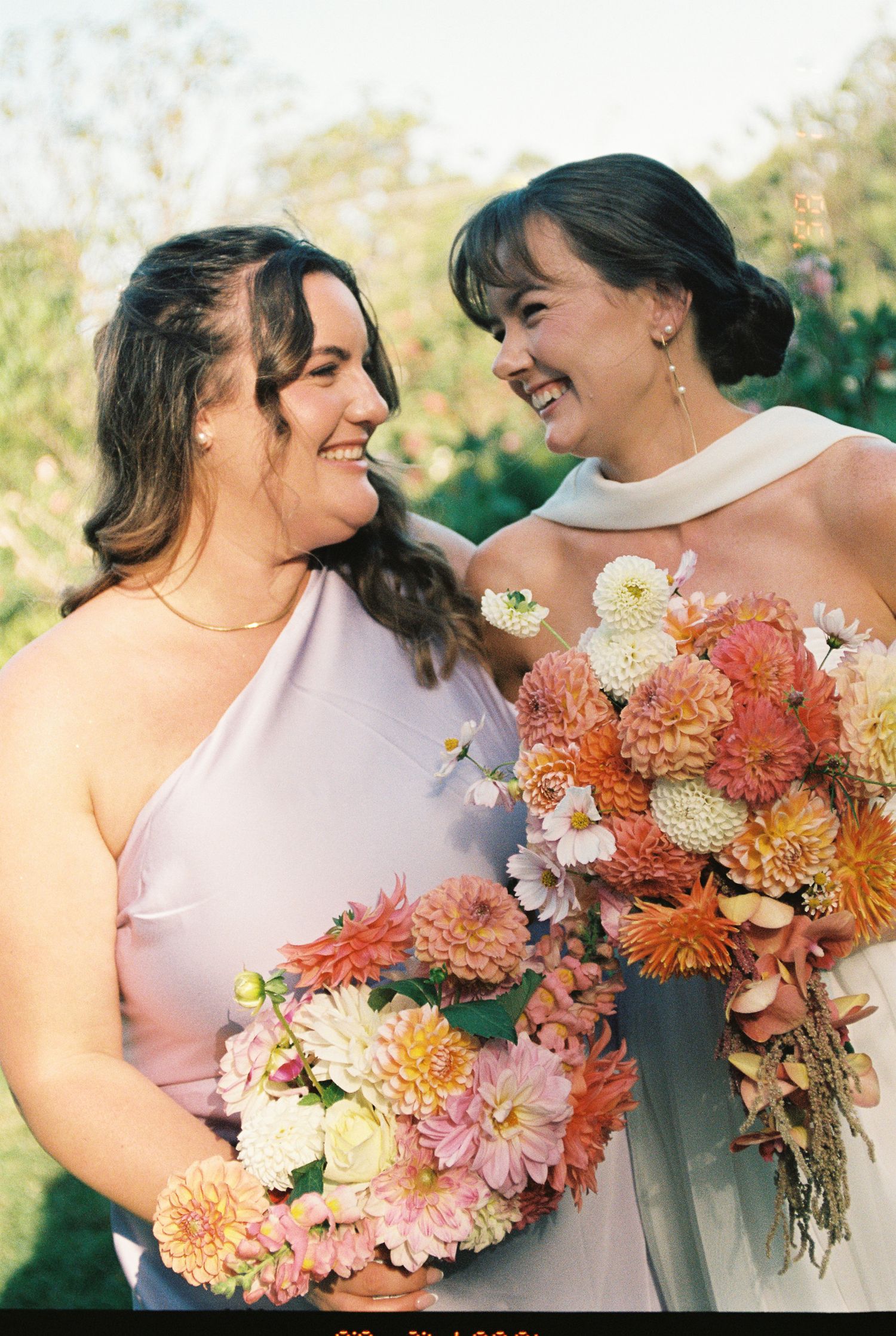 Two smiling women in soft pink and ivory dresses hold colorful bouquets of dahlias and zinnias in an outdoor garden wedding setting.