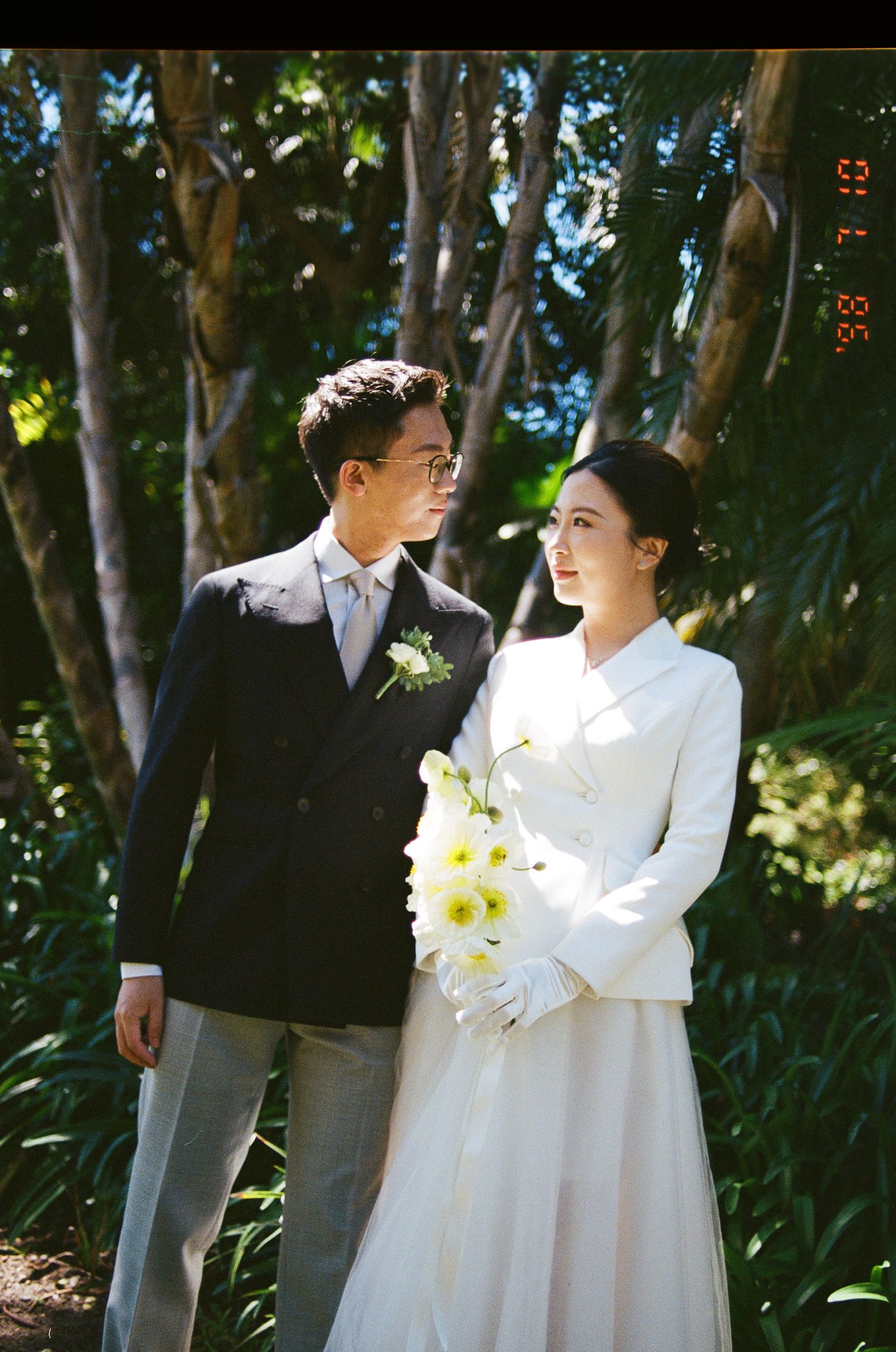 A couple in elegant wedding attire stands in a sunlit garden, the groom in a navy suit and the bride in white holding yellow flowers.