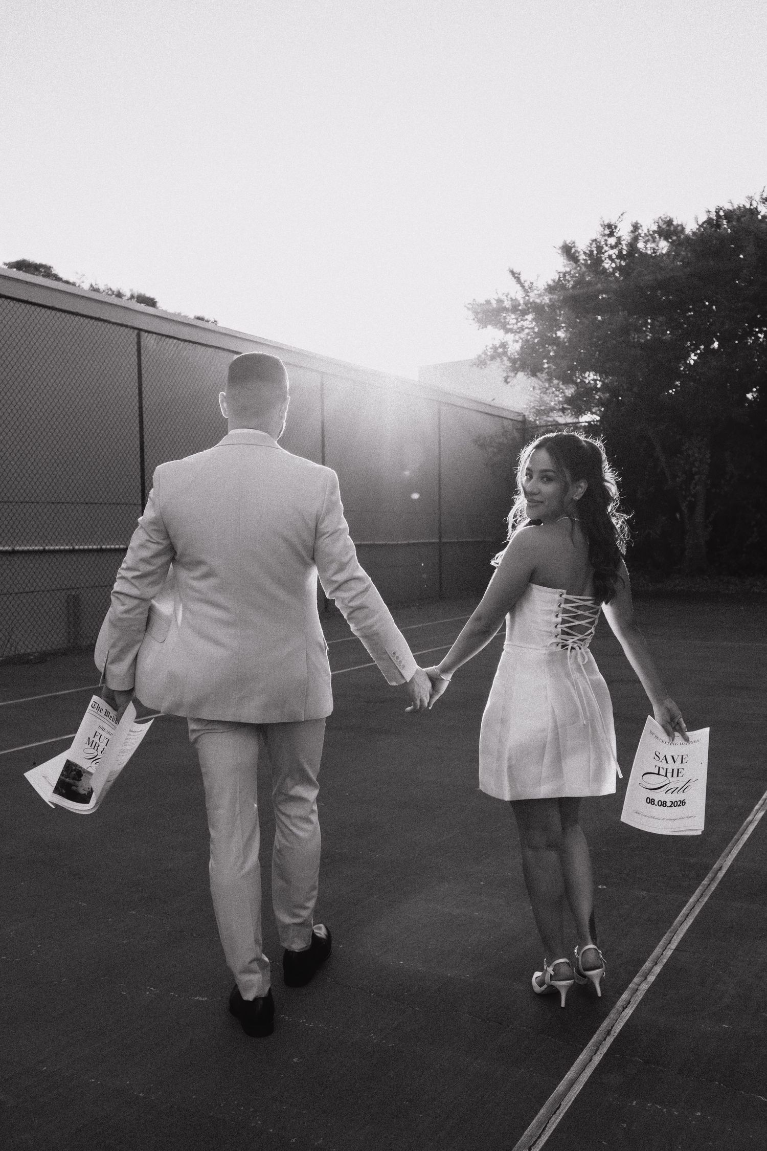 A couple in formal attire walks away holding hands and carrying wine bottles at sunset in black and white.
