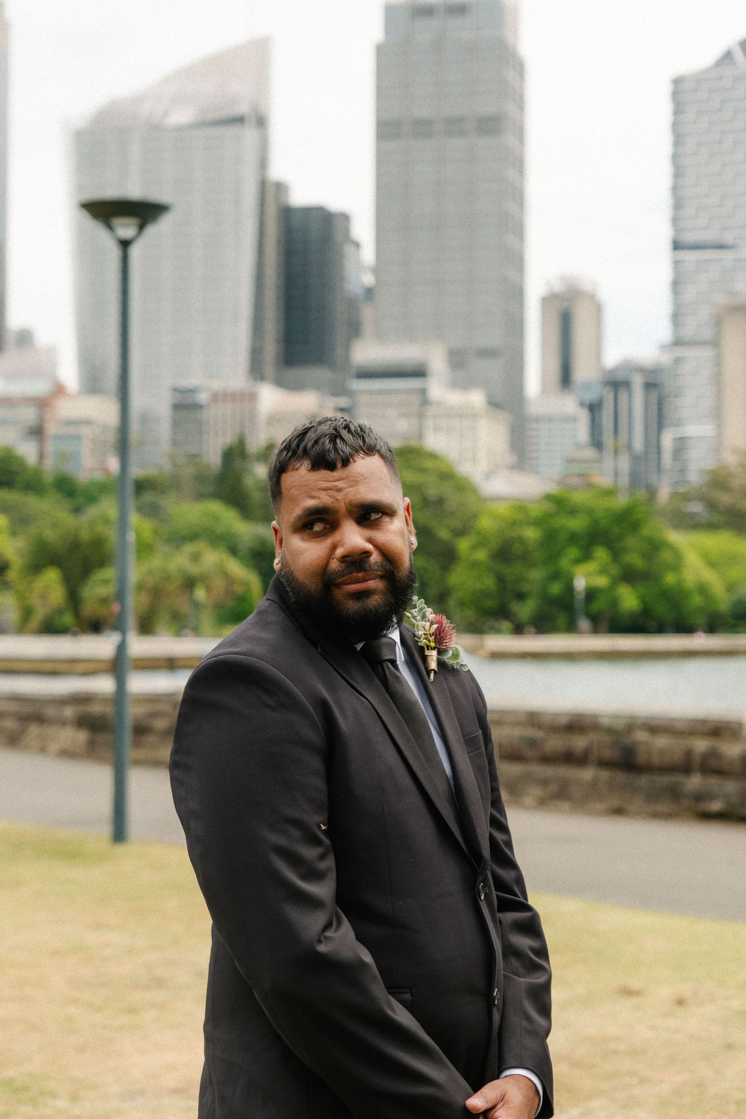 A person in a black suit poses outdoors with a city skyline and park setting in the background.