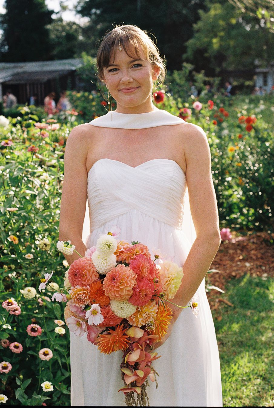 Bride in white strapless gown holding coral dahlia bouquet, posing in lush garden with blooming flowers.