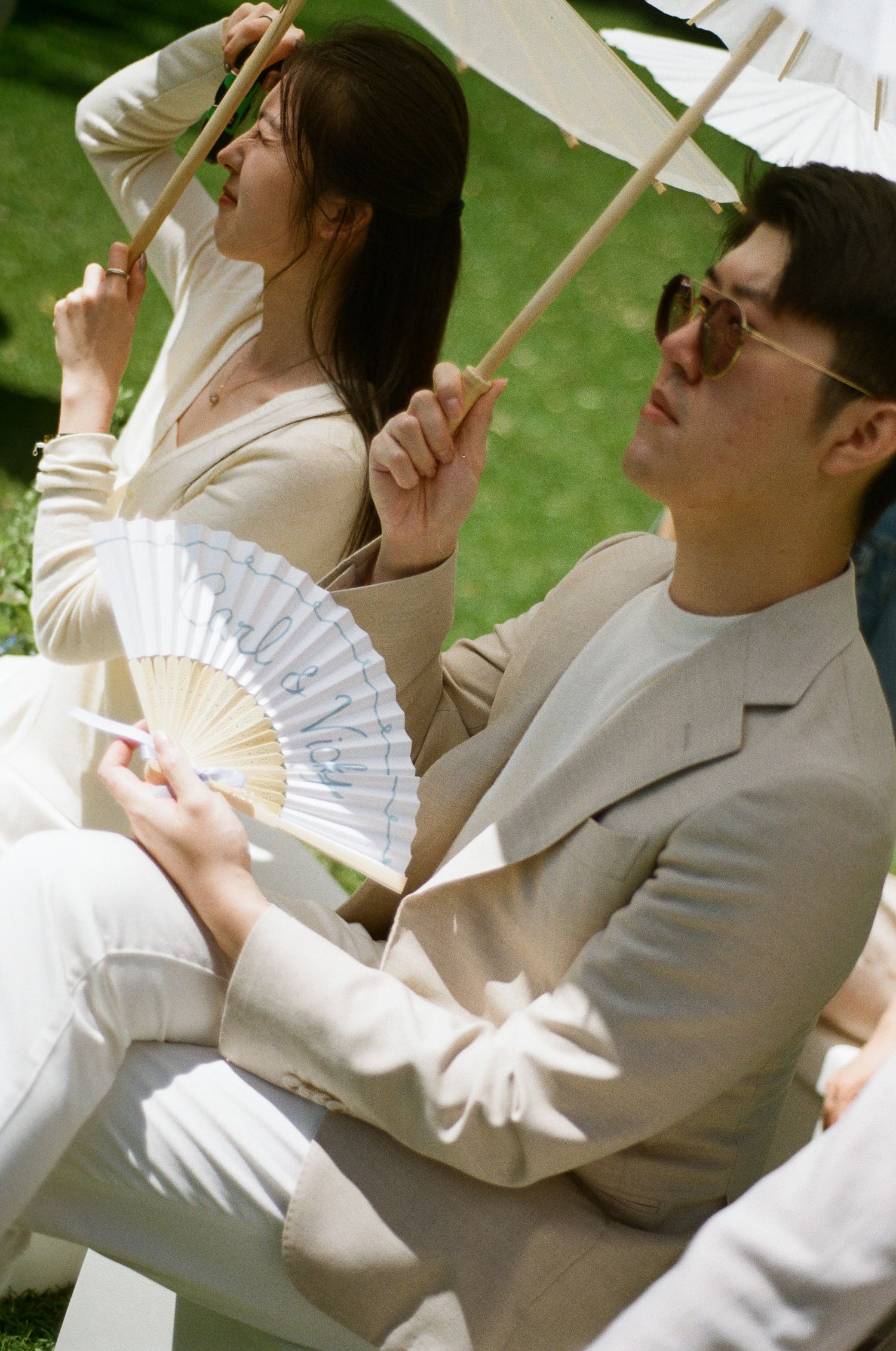 Two guests in cream and beige outfits hold white paper parasols and a folding fan while sitting on the grass at an outdoor event.