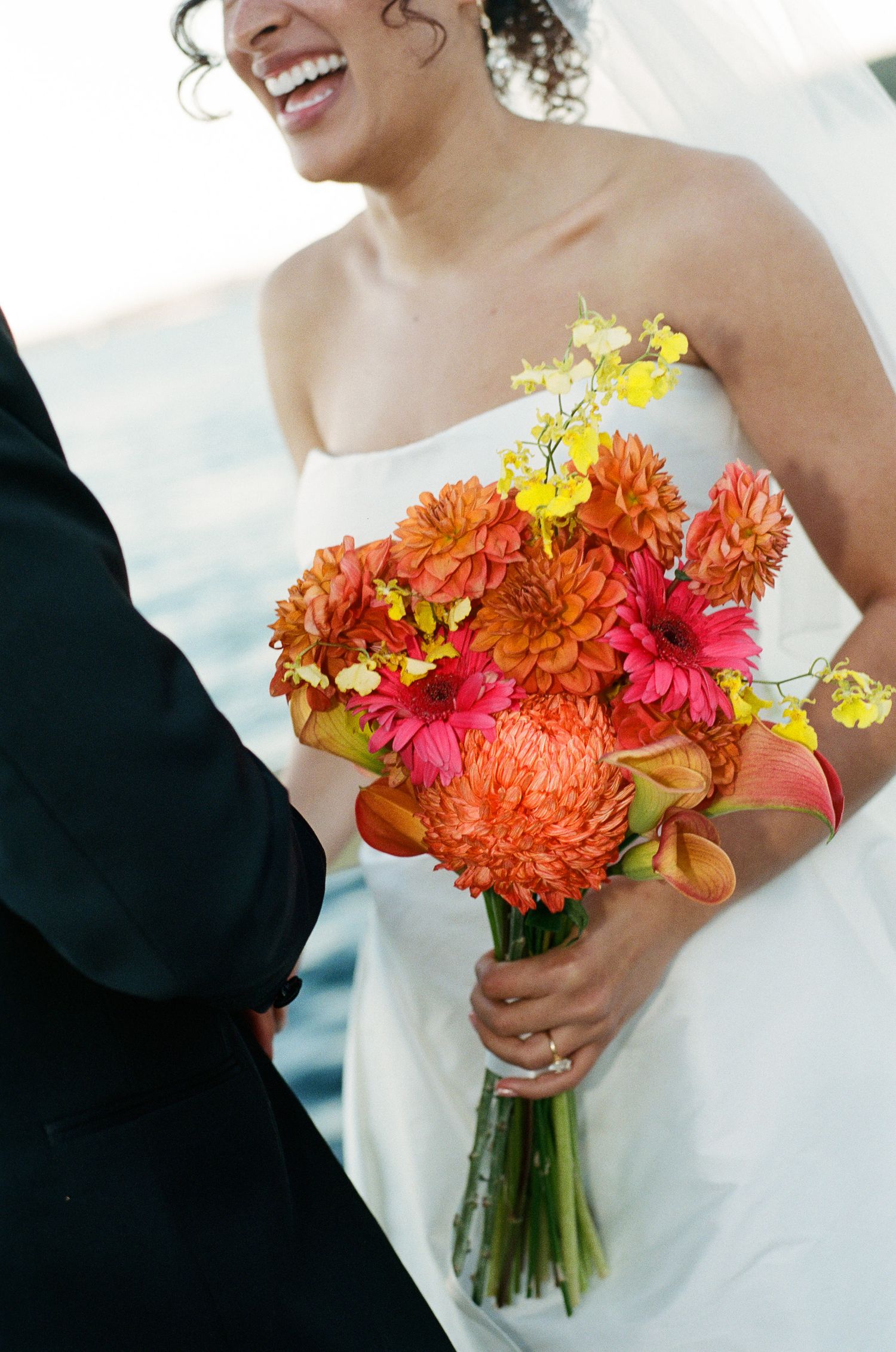 Smiling bride in a strapless white gown holds a vibrant orange, pink and yellow wedding bouquet beside a groom in a dark suit.