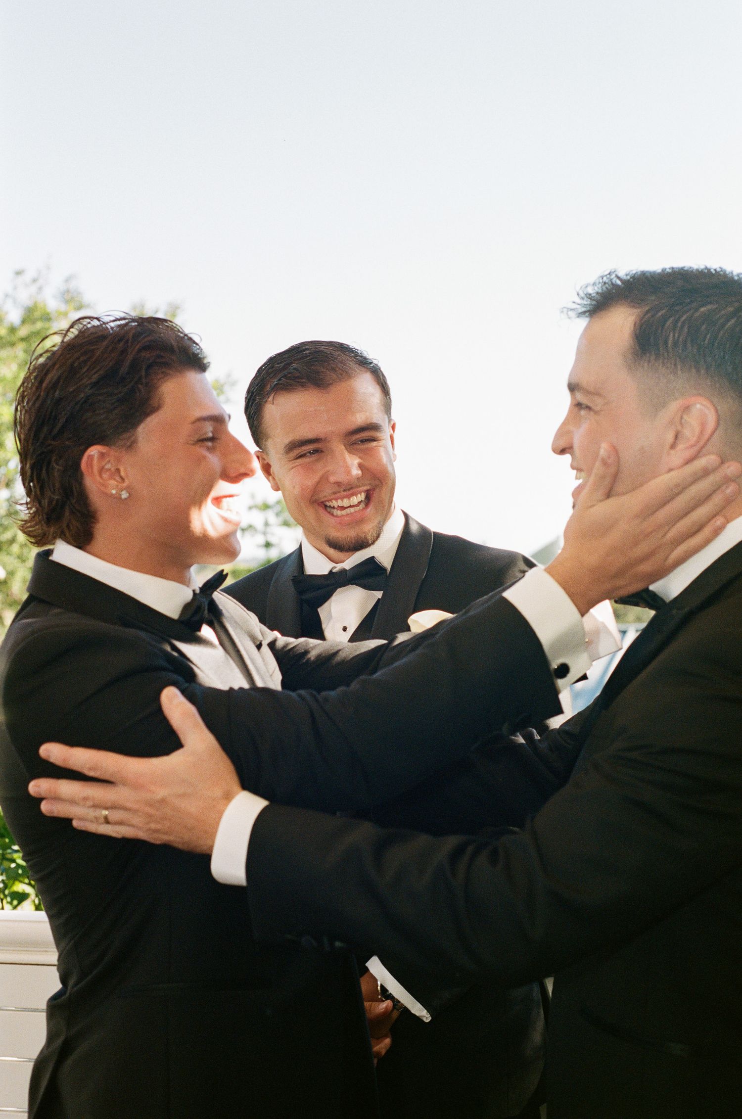 Three men in black tuxedos and bow ties share a joyful embrace at a wedding, laughing as one cups another's face.