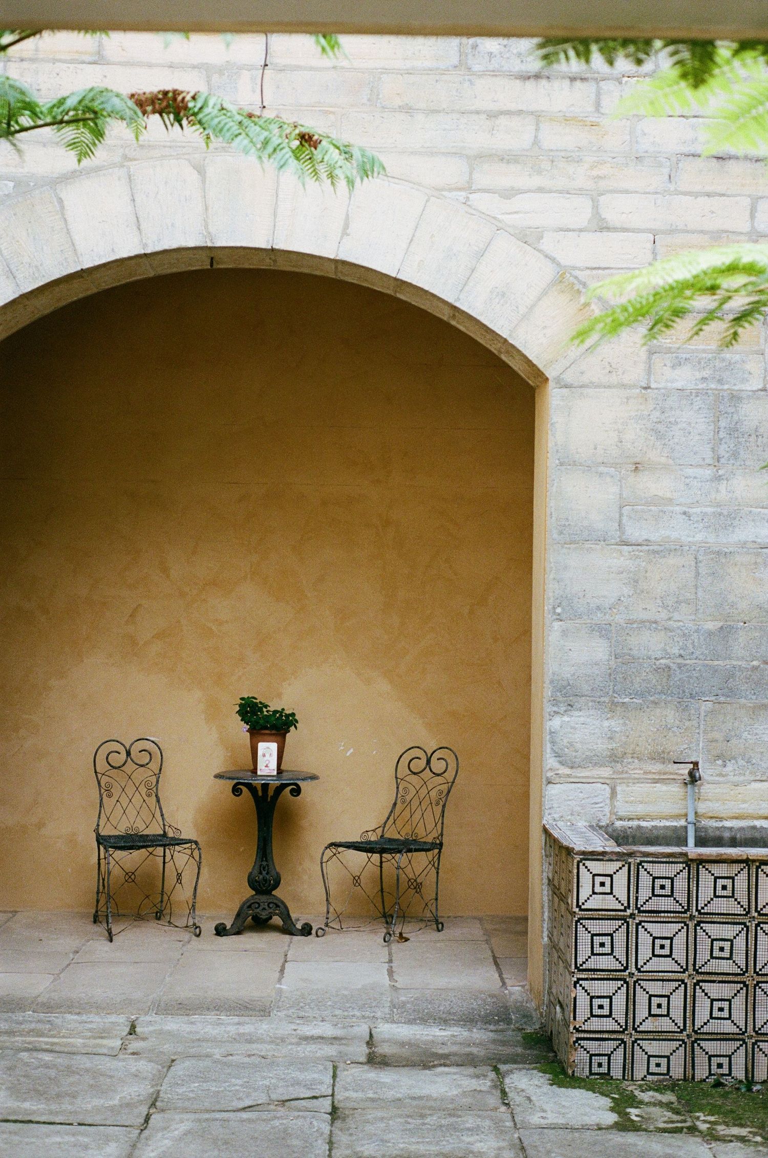 Stone archways with ornate metal chairs and wall sconces create a serene outdoor seating area in a Mediterranean courtyard.