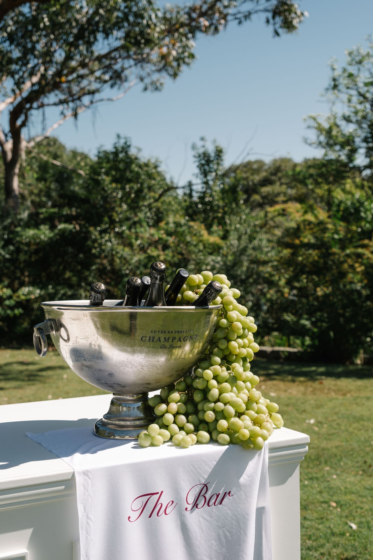 A silver champagne bowl filled with bottles sits on a white-clothed table labeled 'The Bar' with fresh green grapes.