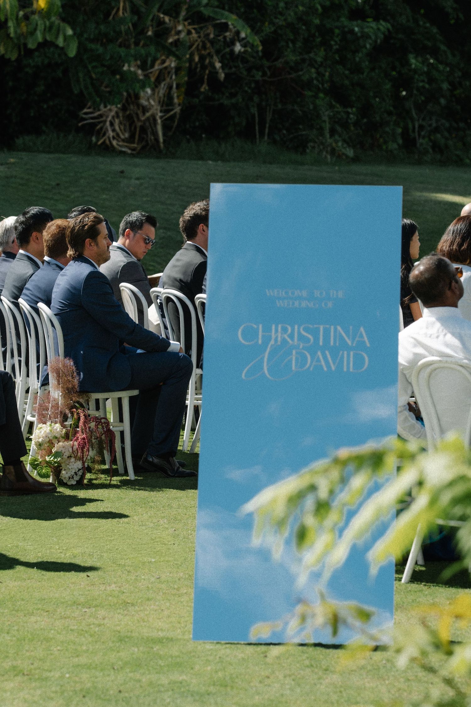 Wedding guests seated outdoors on white chairs with blue signage displayed on a sunny lawn.
