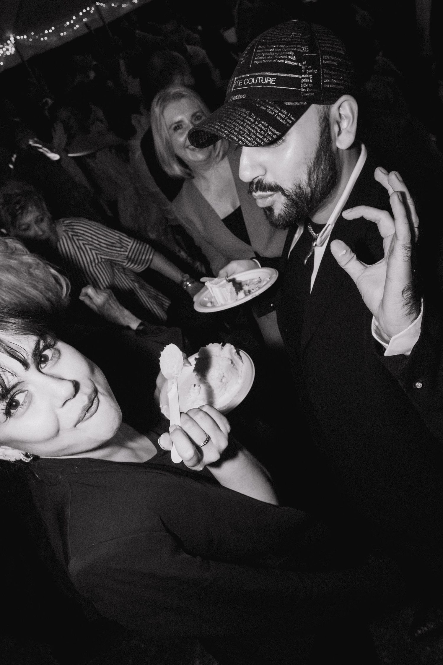 Black and white party photo of a bearded man in a cap making an OK gesture beside a smiling woman holding cake, with guests behind.