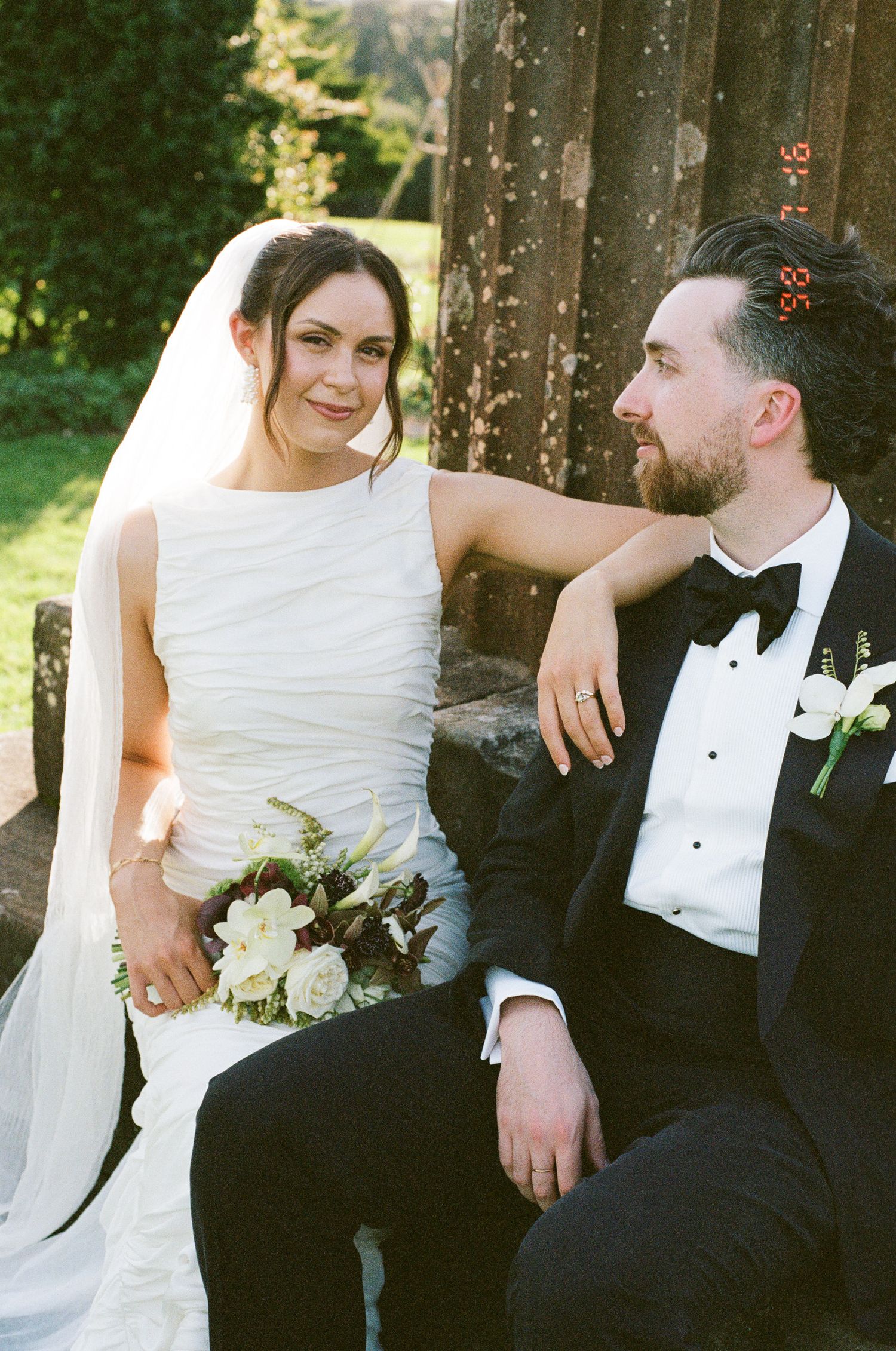 Smiling bride in a white sleeveless gown and veil holds a white floral bouquet while leaning on a groom in a black tuxedo and bow tie.