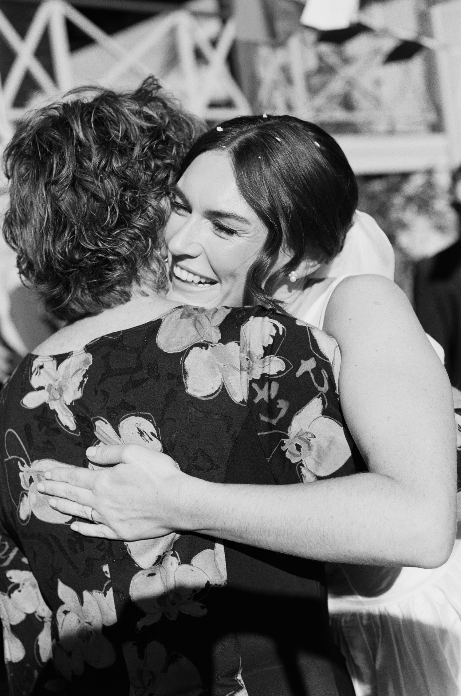 Black and white photo of a smiling bride in a white dress warmly hugging a guest in a floral top at an outdoor wedding.