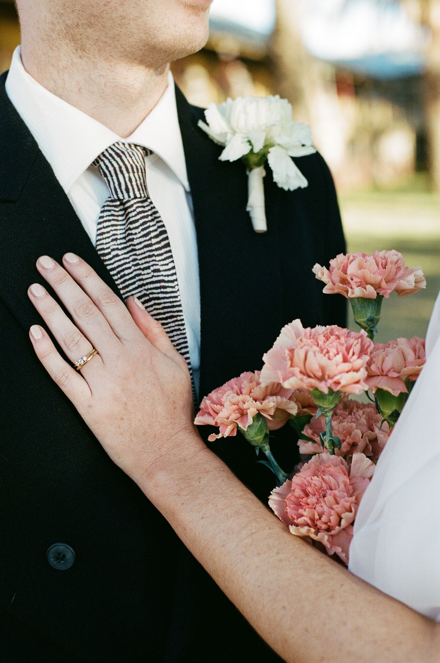 Bride's hand with a gold wedding ring rests on a groom's black suit beside a pink carnation bouquet and white boutonniere.