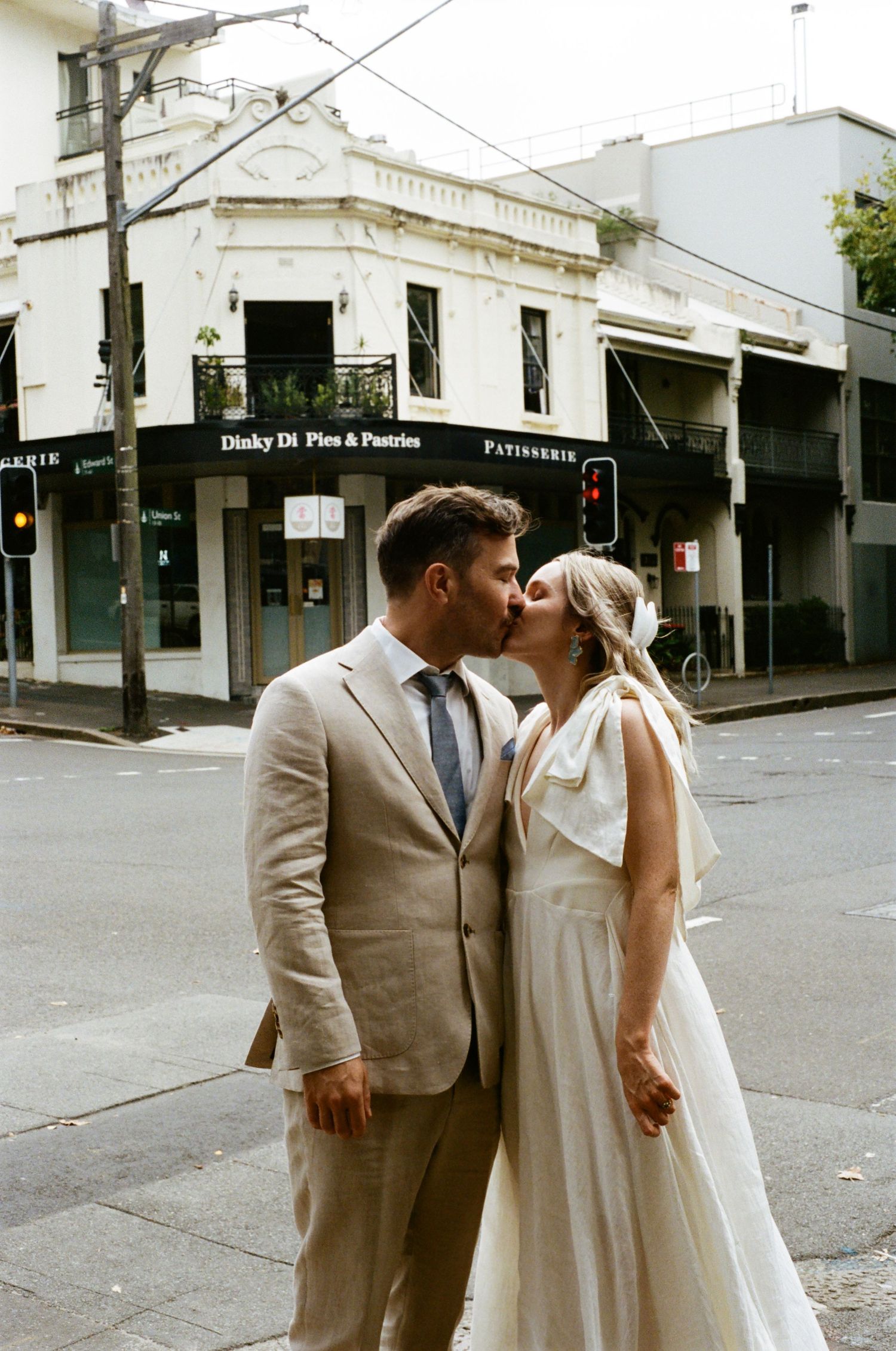 A couple shares a romantic kiss on a city street corner in front of a vintage storefront building.