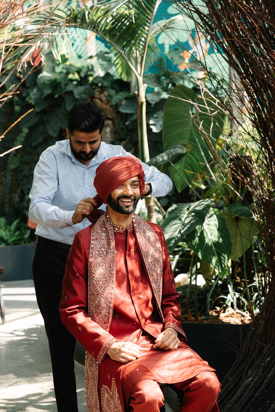A groom in a traditional red embroidered Indian wedding attire gets ready while seated in a garden setting.
