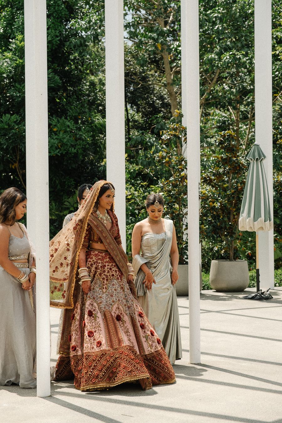 A wedding party in elegant attire stands between white pillars in a garden setting, featuring a bride in an ornate brown lehenga.