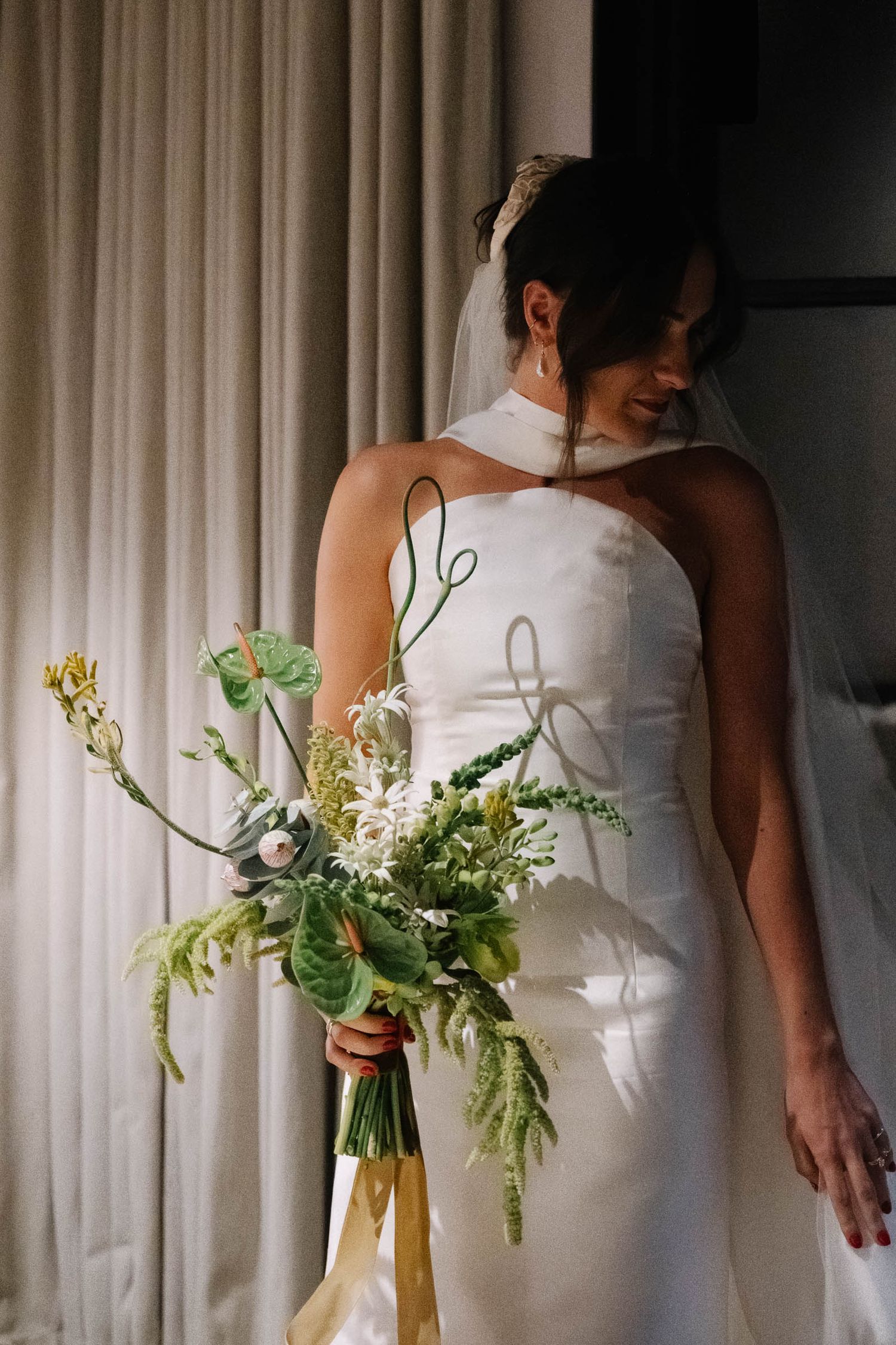 Row of bridesmaids in white dresses holding green and white floral bouquets against light curtain backdrop.