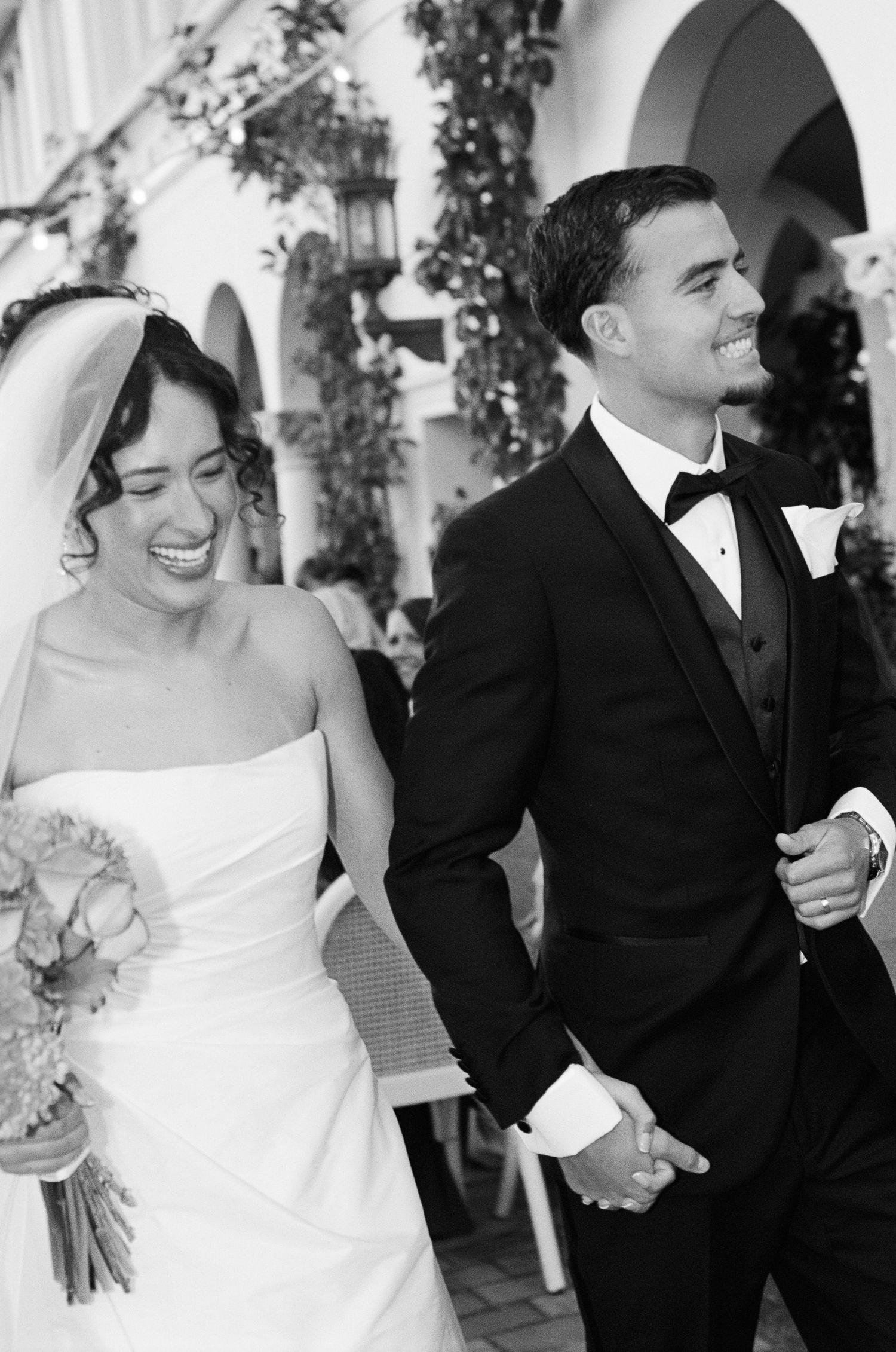 Black and white photo of a laughing bride in a strapless gown and veil holding a bouquet, walking hand in hand with her groom in a tuxedo.