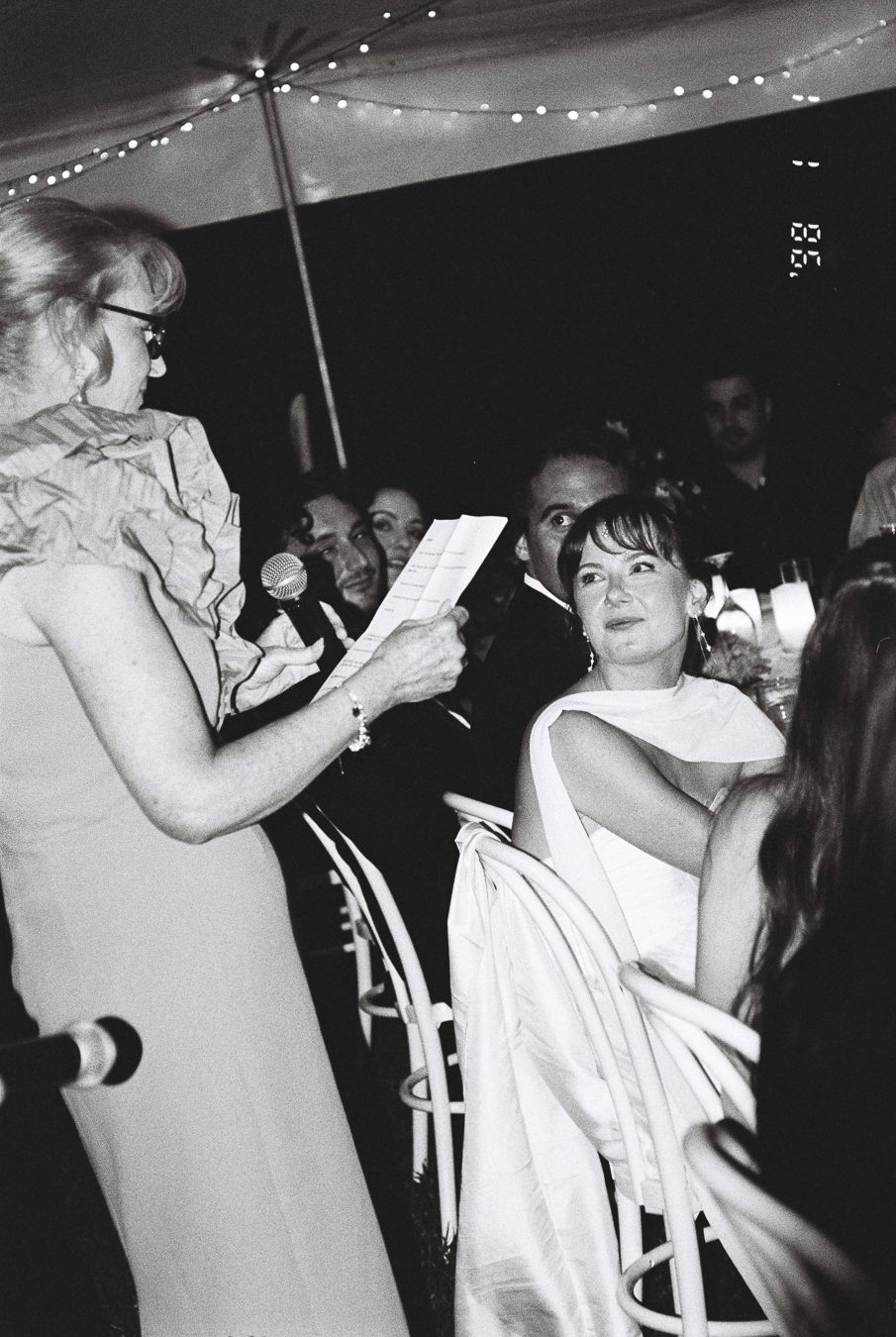 Black and white photo of women at a formal event, one reading a paper while another in white dress listens attentively.
