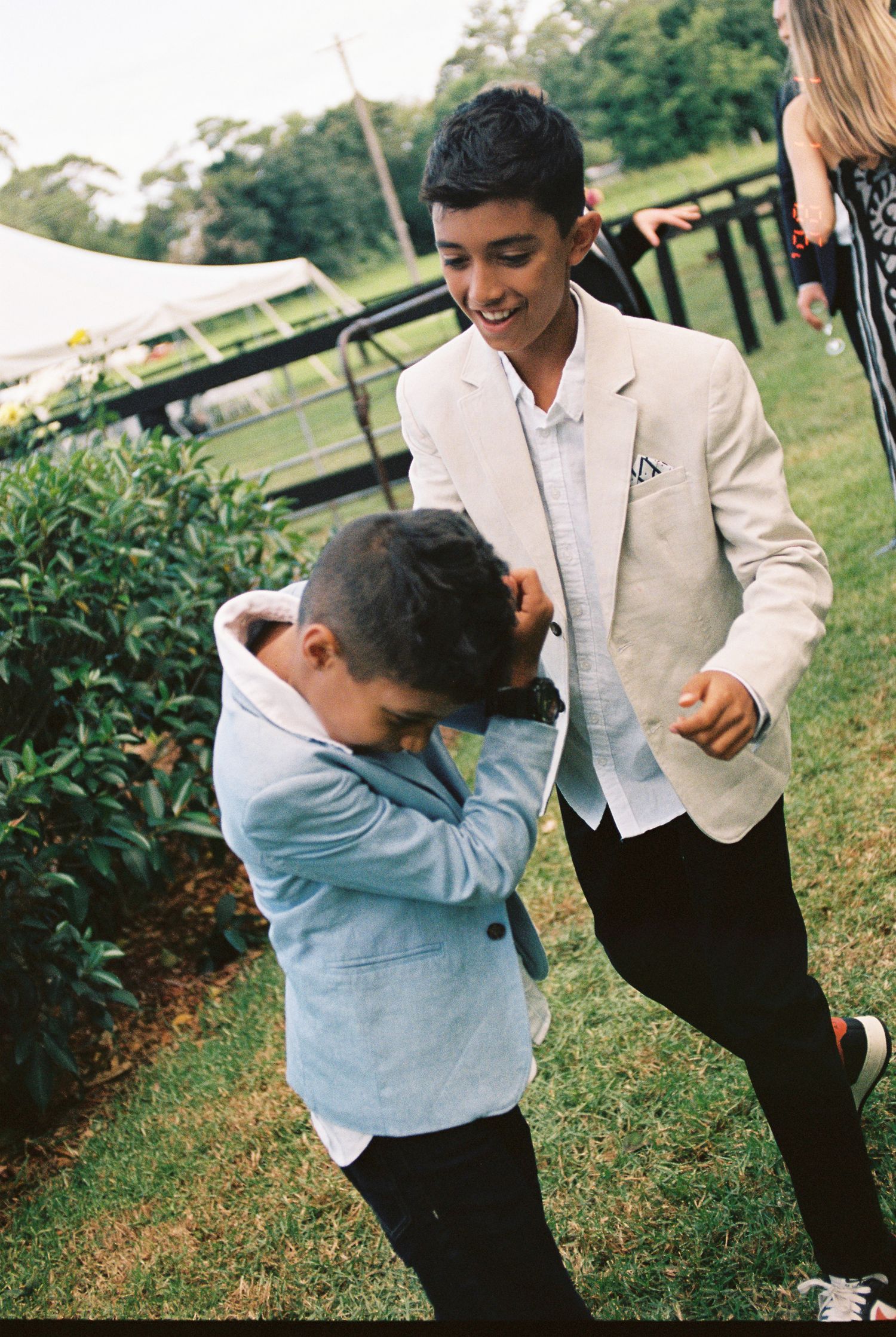Two boys in blazers play on a grassy lawn at an outdoor event, one laughing in a cream jacket while the other ducks in a light blue jacket.