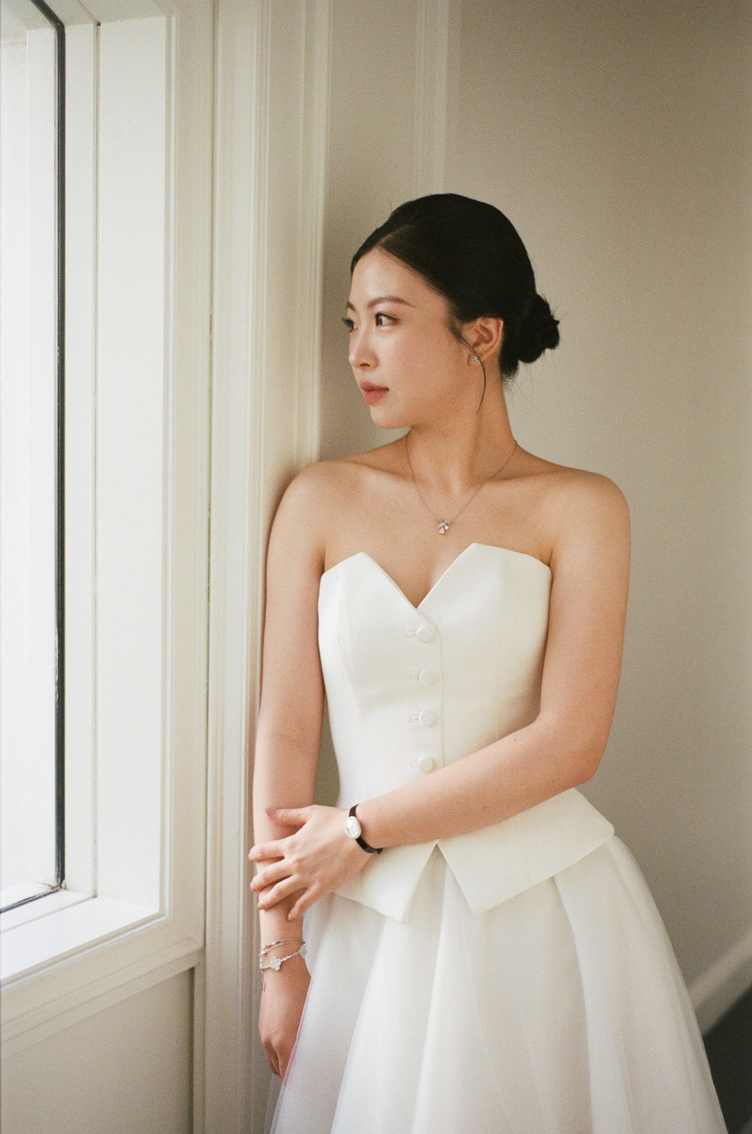 Woman with dark hair in an updo wearing a white strapless bustier top and tulle skirt, standing beside a window in soft natural light.