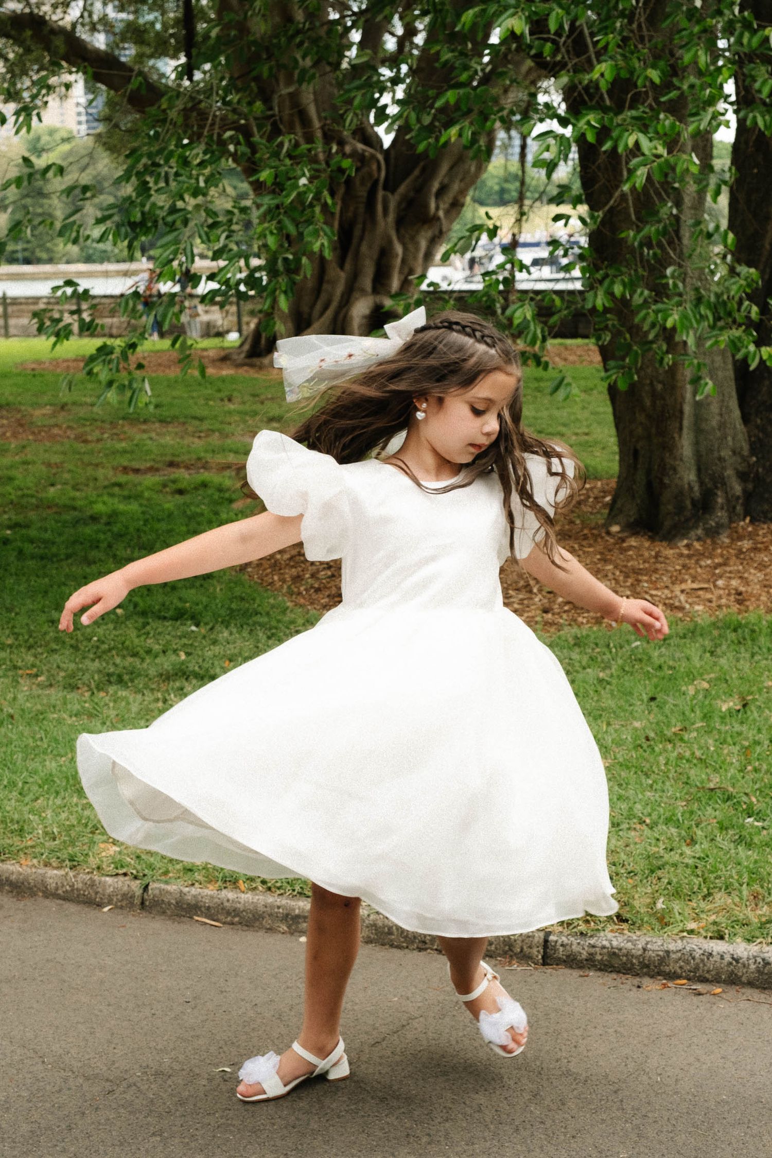 A young dancer in a flowing white dress twirls gracefully on a paved path near trees in a park setting.