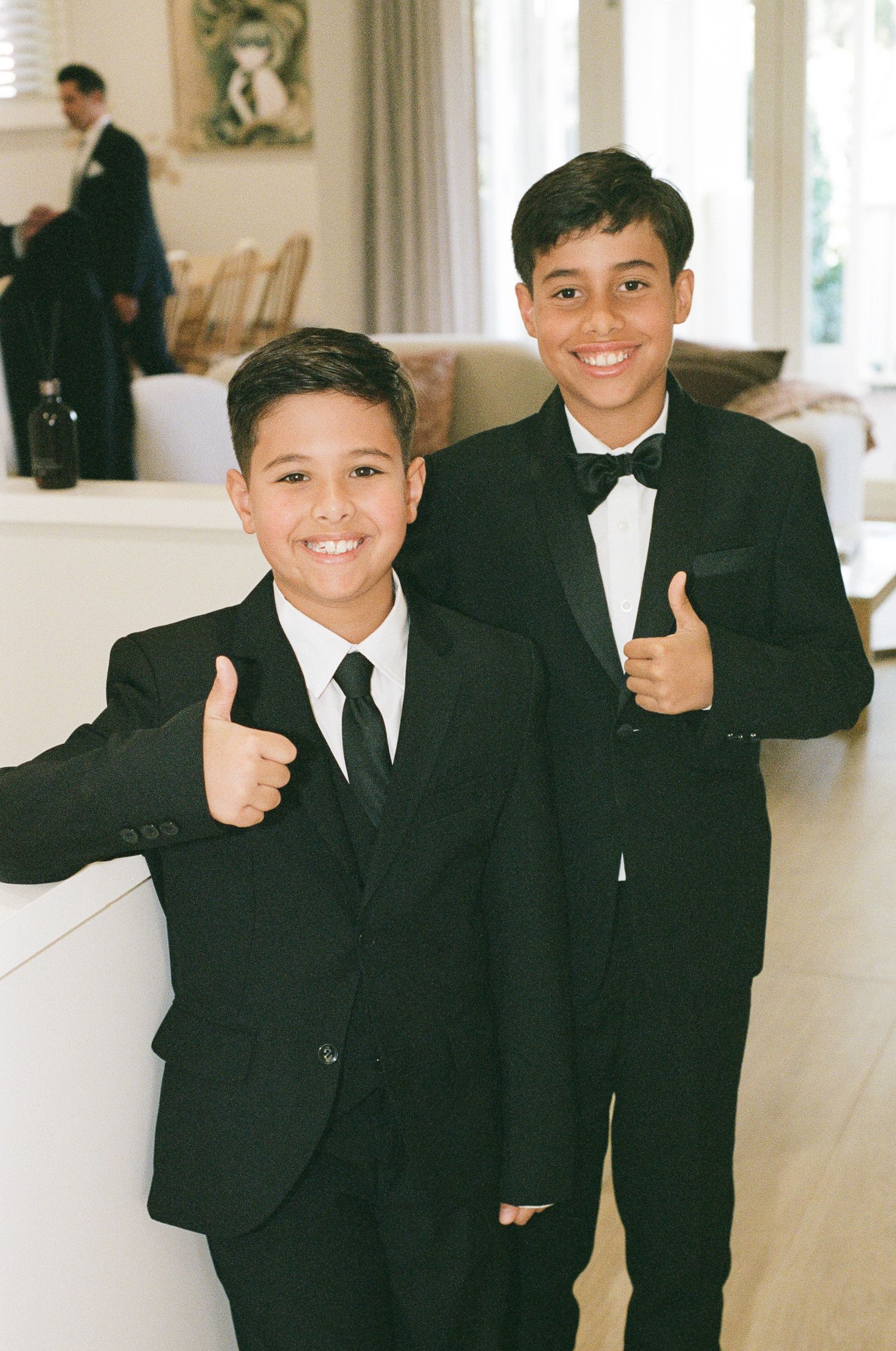 Two smiling boys in black tuxedos and bow ties give thumbs up in a bright room at a wedding celebration.