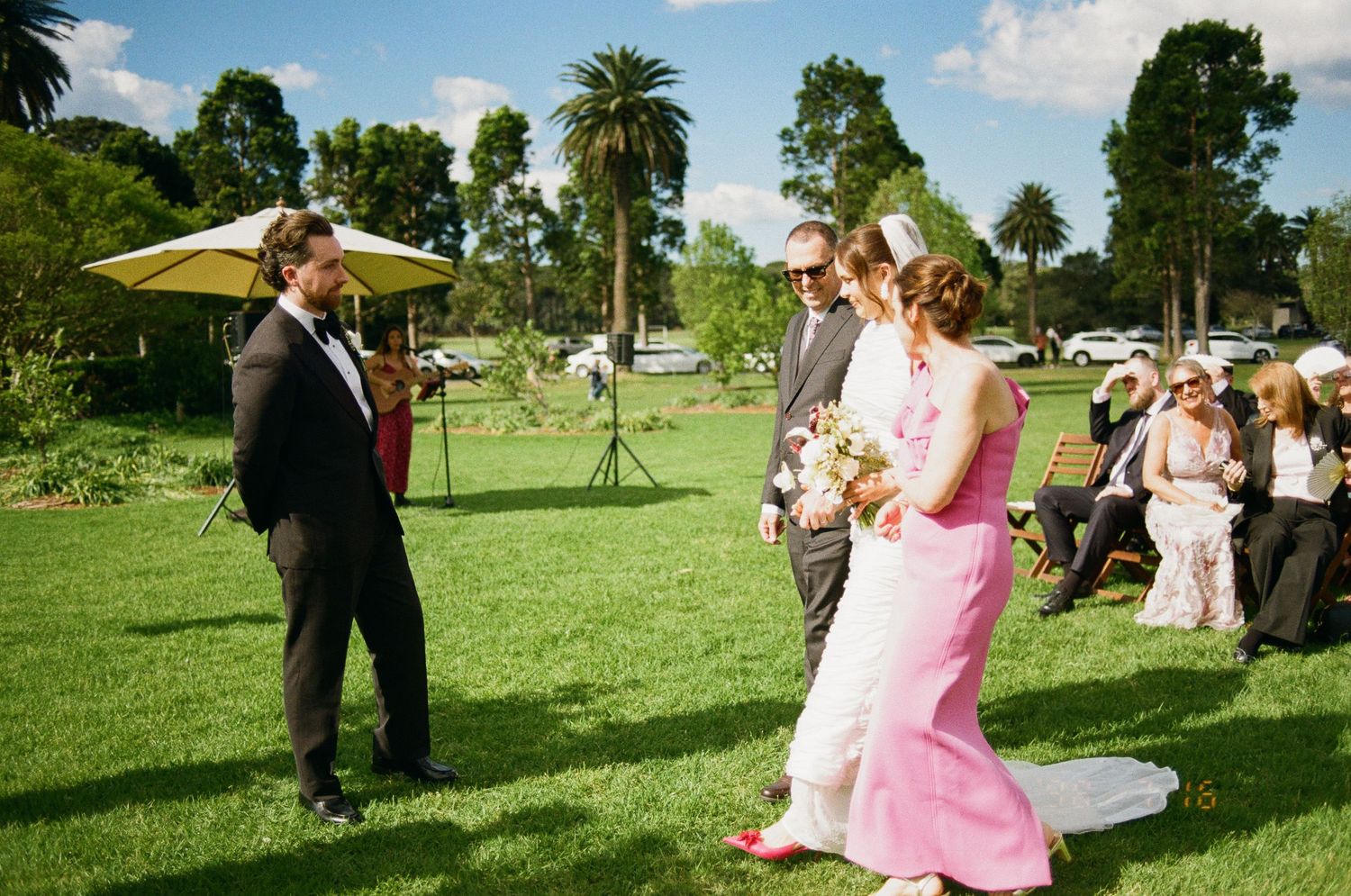 Bride in a pink gown holding a bouquet walks down the aisle at an outdoor garden wedding as guests watch from the lawn.