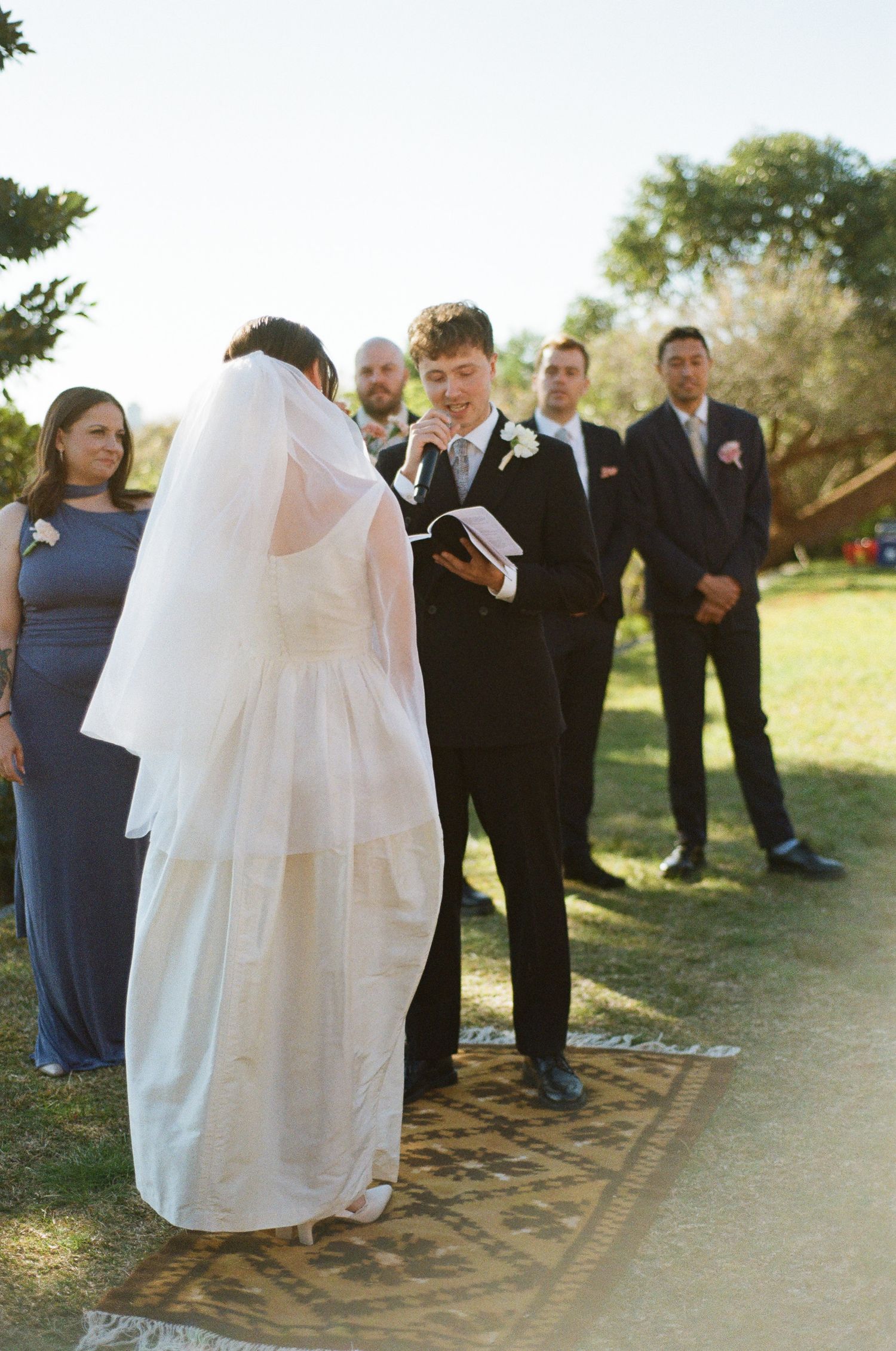 Groom in a dark suit reads his vows from a book as the bride in a white dress and veil faces him on a rug outdoors.