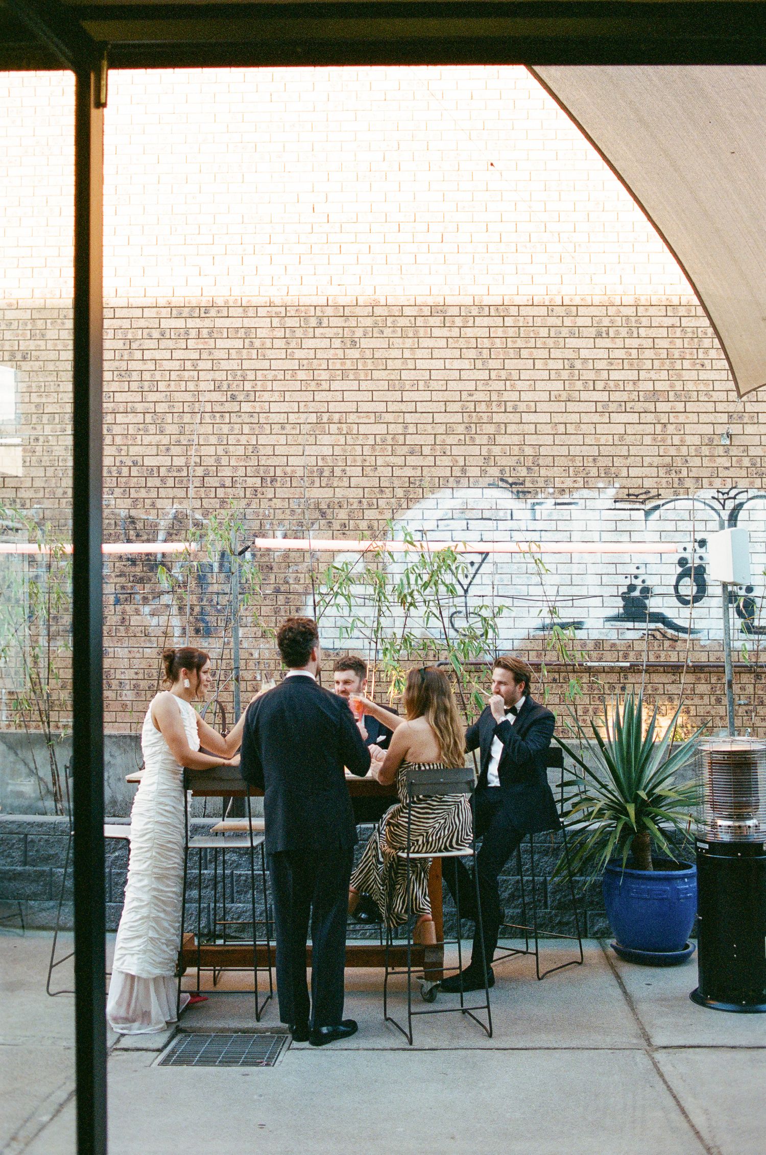 Wedding guests in formal attire chat around a high-top table on a brick-walled rooftop patio with graffiti, a potted agave and a patio heate