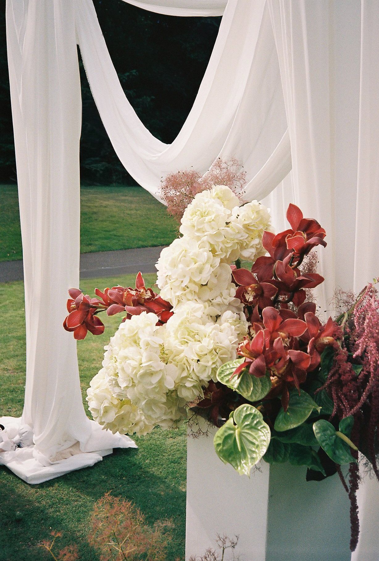 White fabric draping frames elegant floral arrangements with white hydrangeas and burgundy flowers at an outdoor event.
