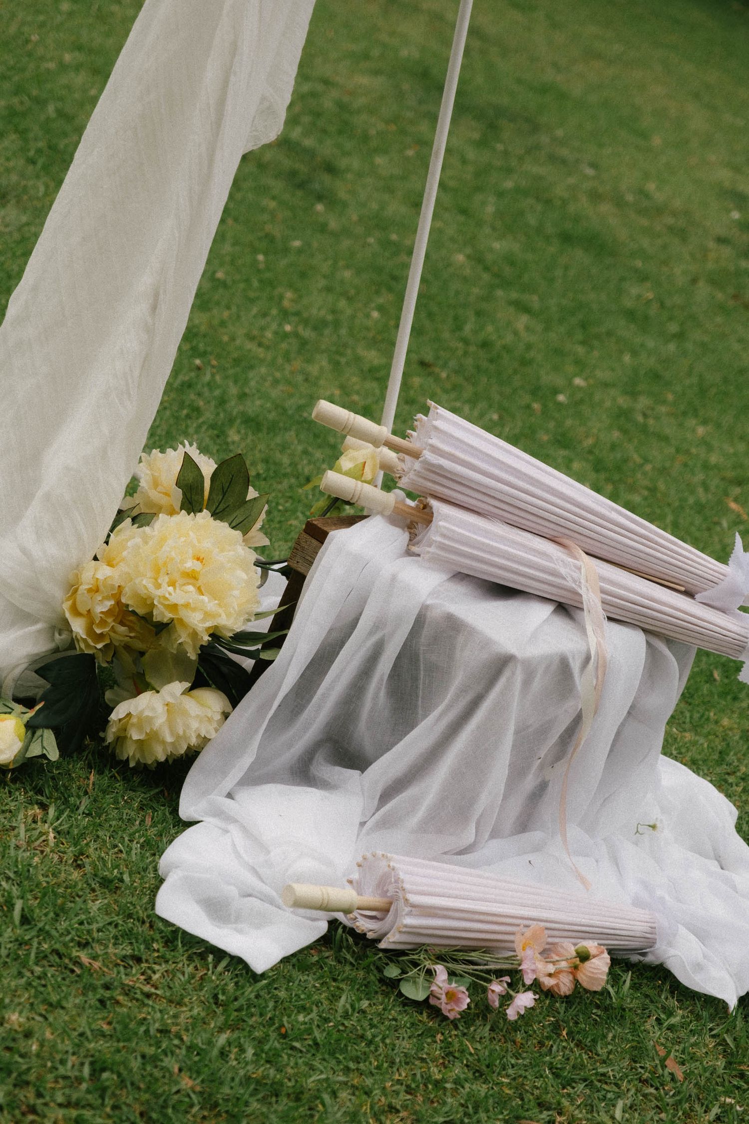 White fabric and floral decorations lay scattered on green grass creating an ethereal outdoor wedding scene.