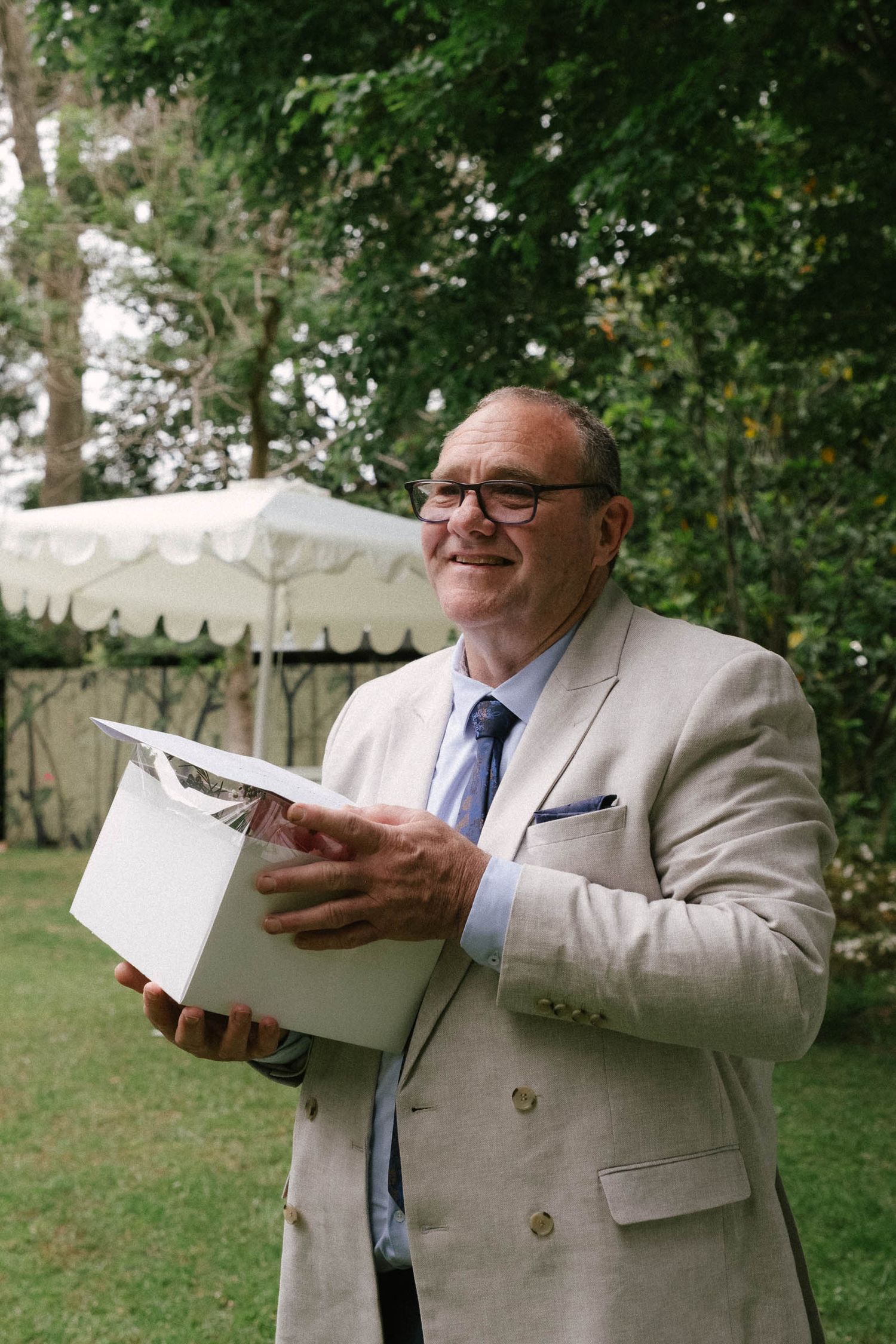 Someone in a light beige suit and glasses holds a white box while standing in a garden setting.
