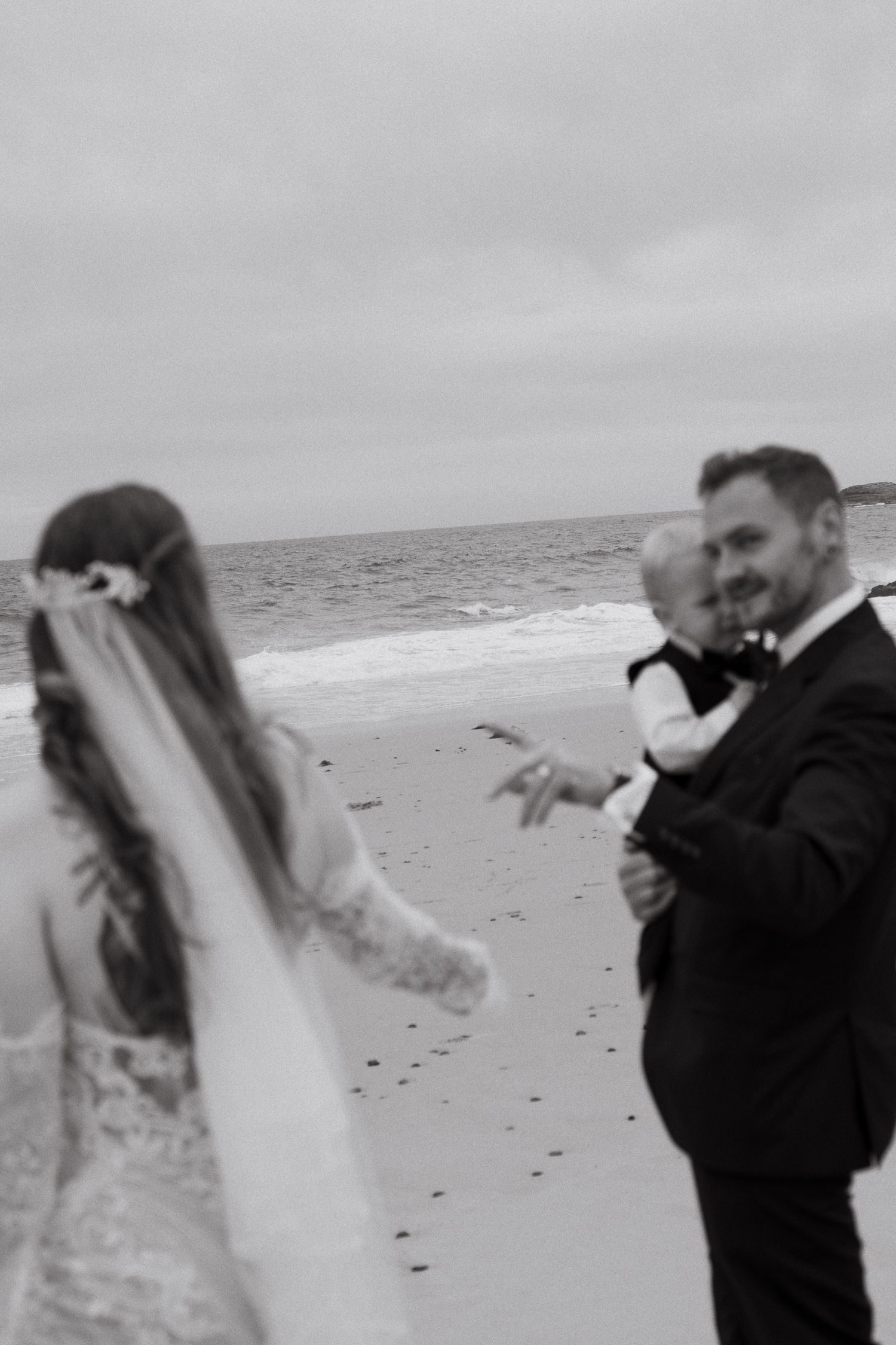 A couple shares a romantic moment on a windy beach with a long veil flowing in the breeze.