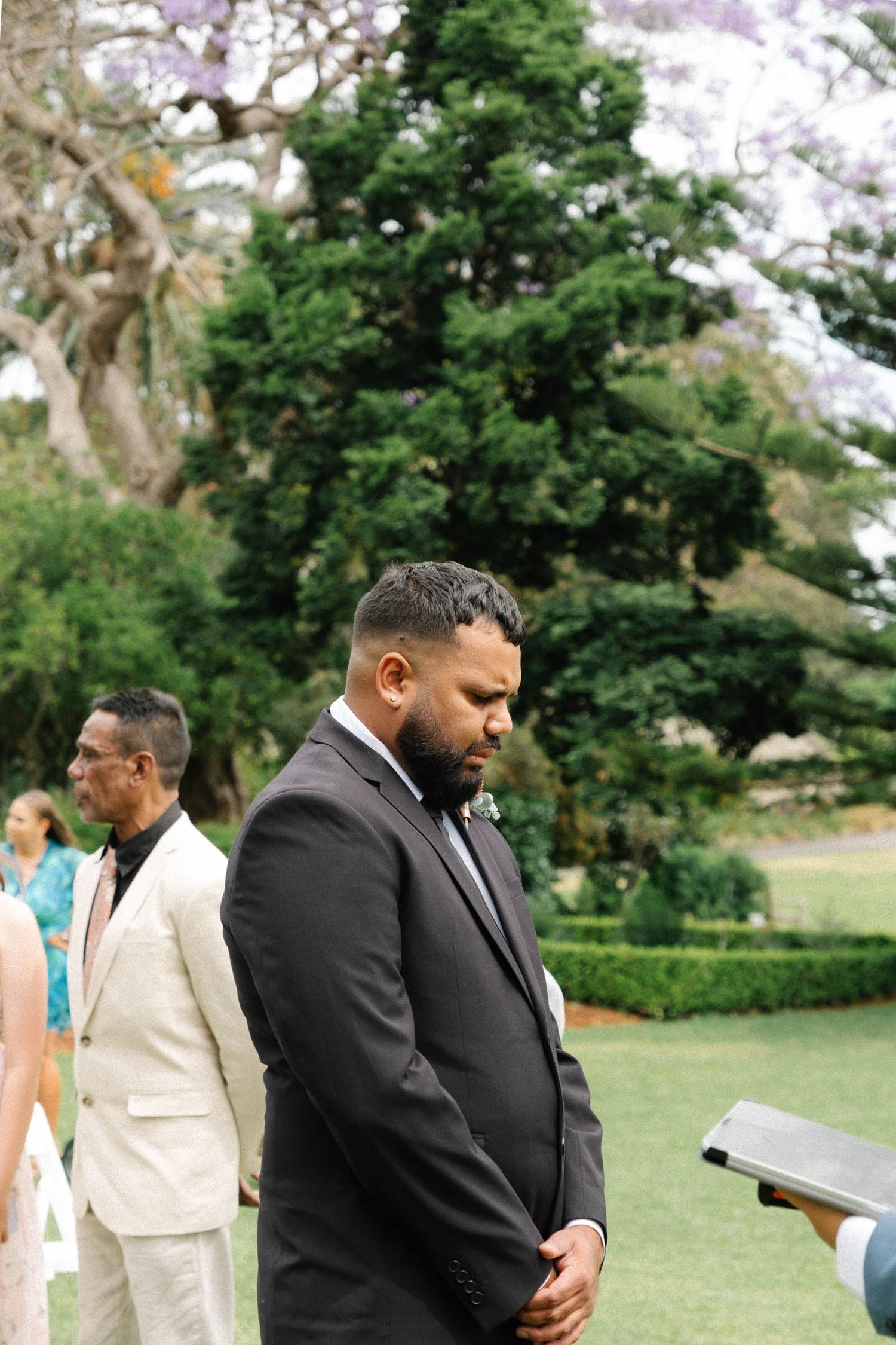 Wedding party members in black and cream colored suits gather together outdoors during a ceremony.