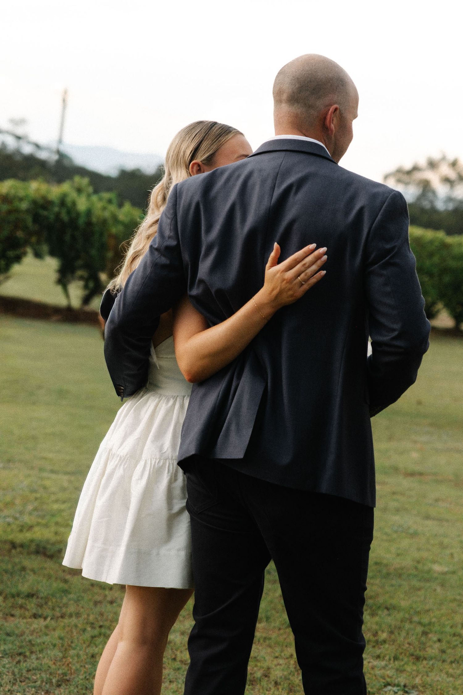 A couple in formal wedding attire embraces while walking away from the camera in a grassy outdoor setting.