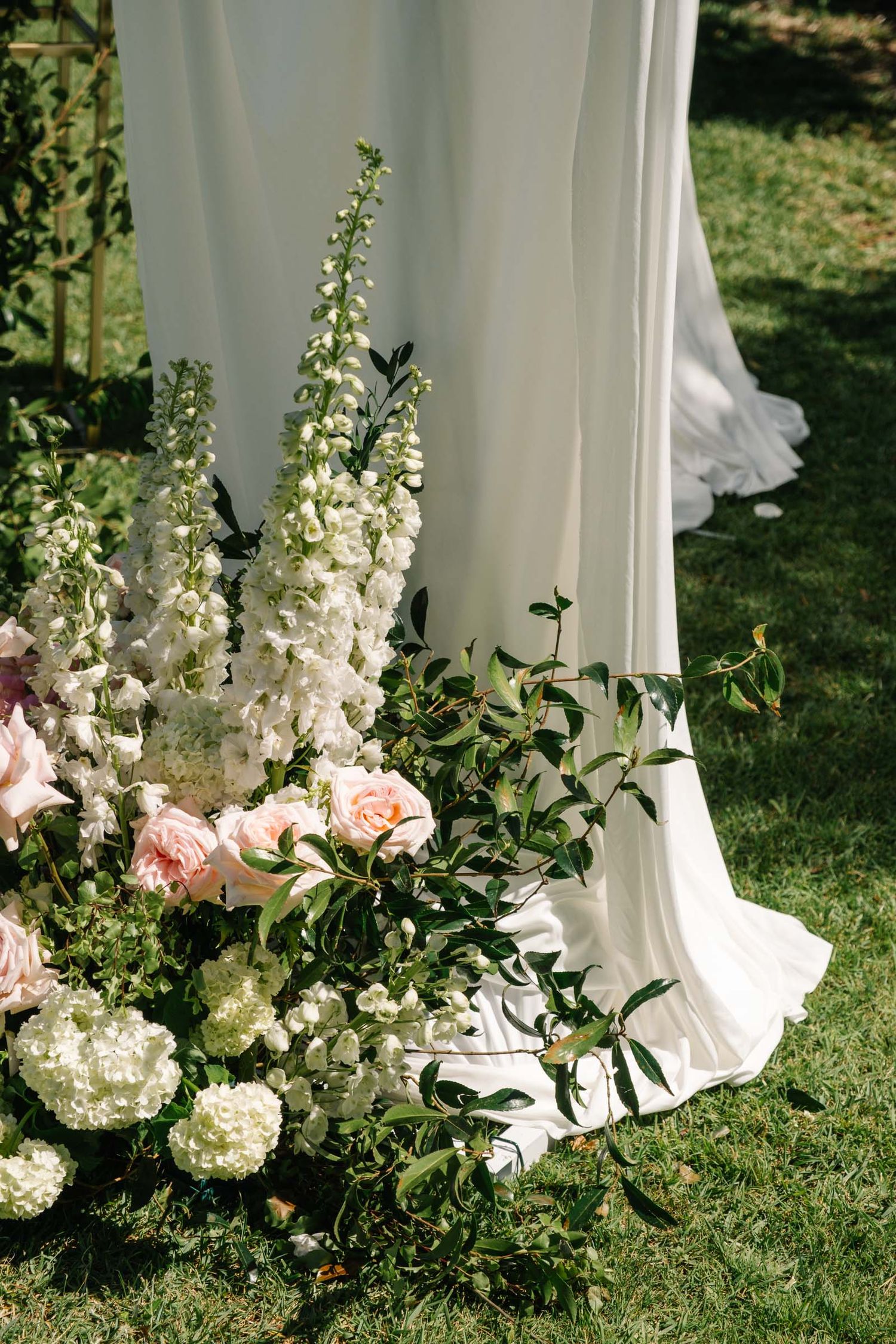 Elegant white floral arrangements with pink roses and greenery line a white fabric backdrop on grass.