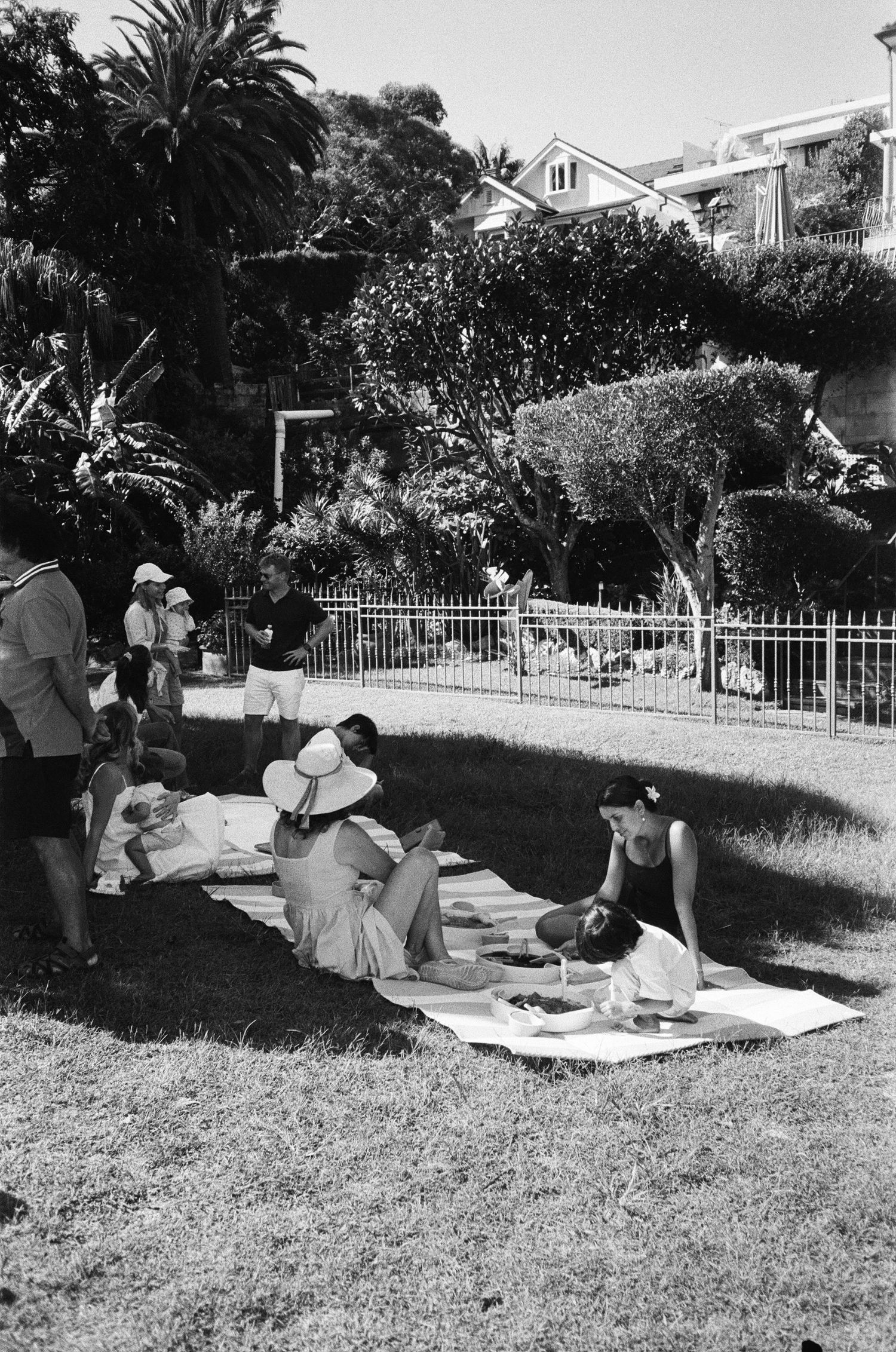 A group of people relax on blankets while having a picnic on a grassy hillside in black and white.