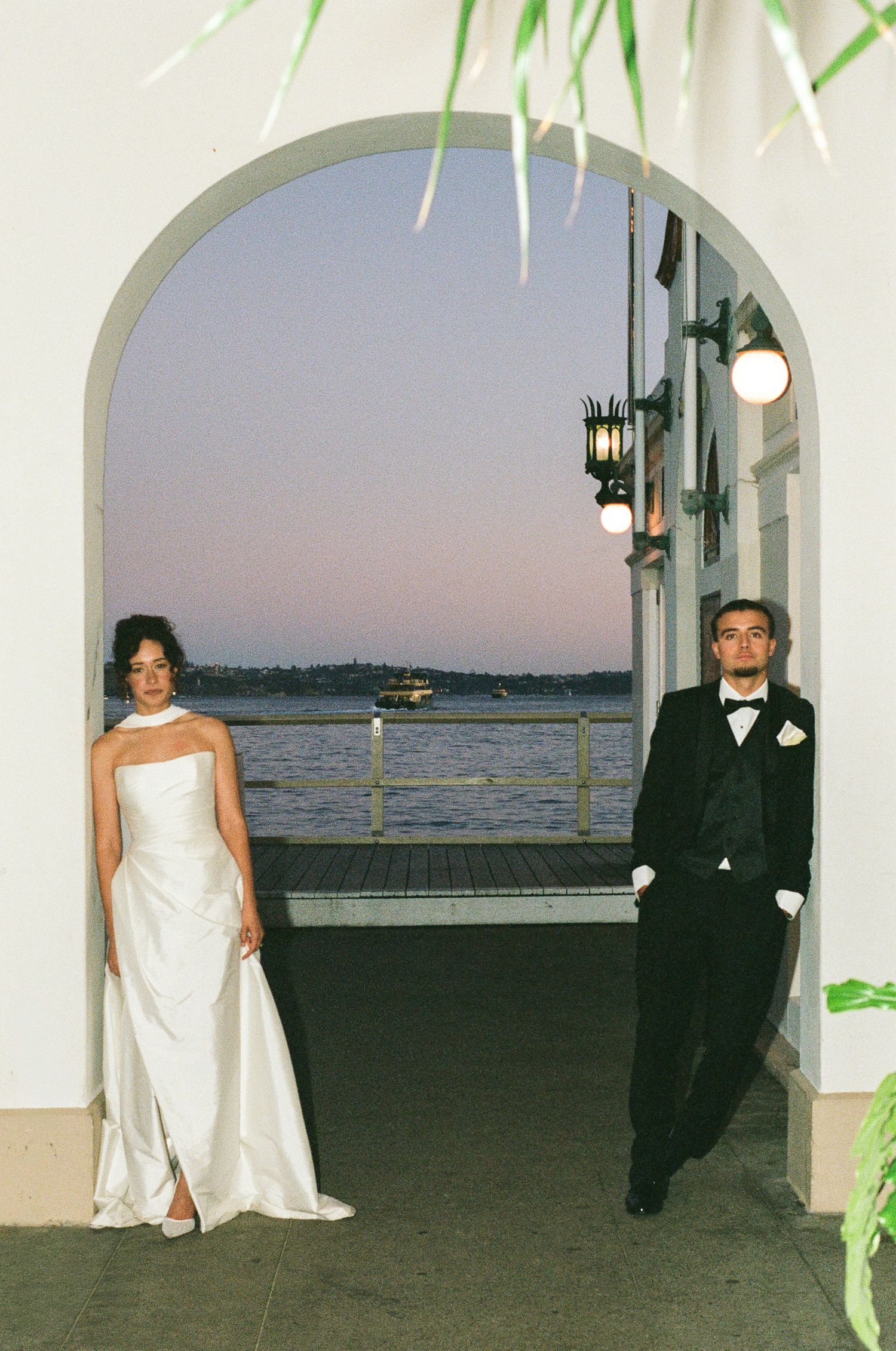 Bride in a white strapless gown and groom in a black tuxedo pose under a white archway overlooking the water at dusk.