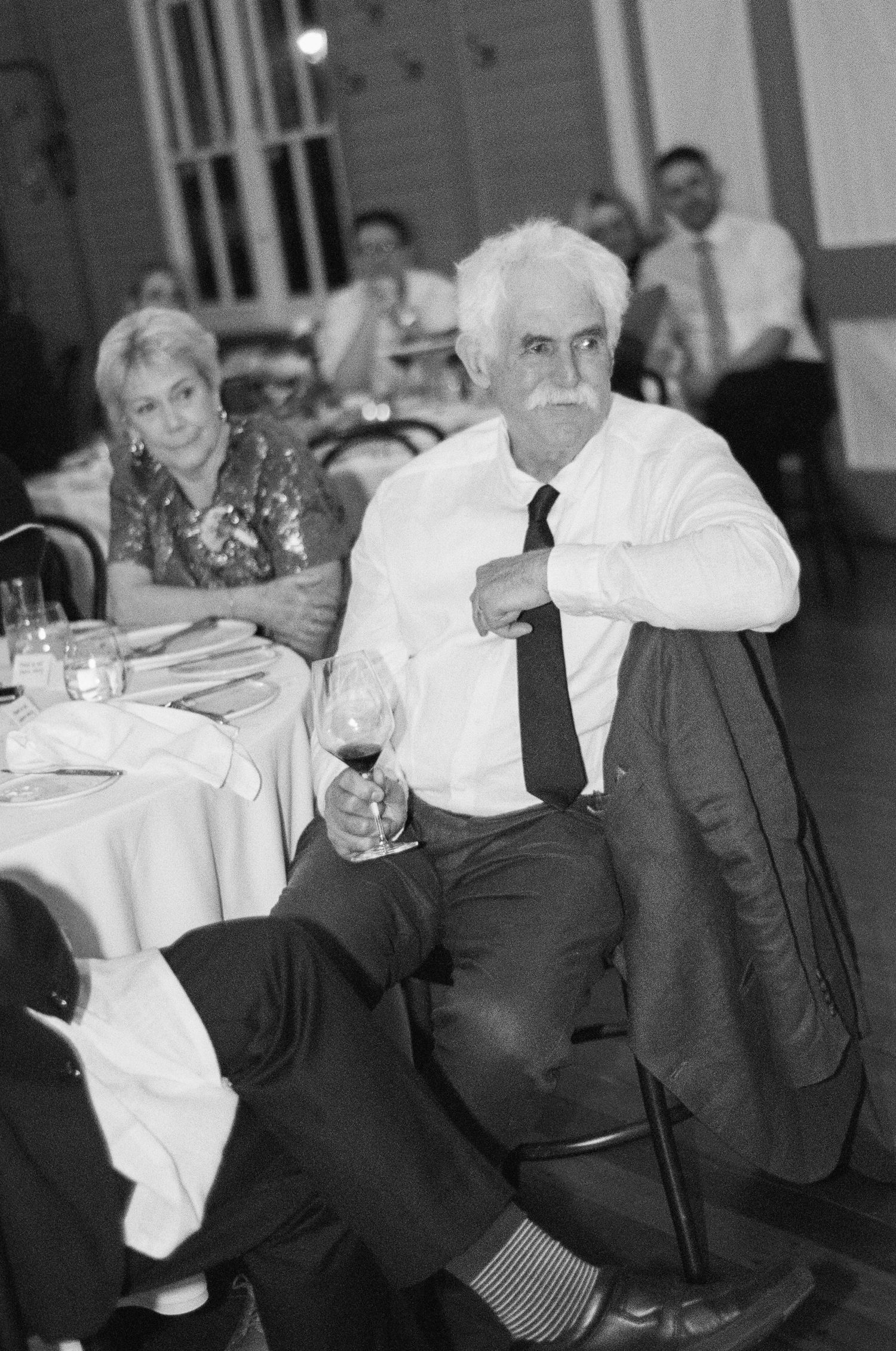 Black and white photo of a white-haired man with a mustache holding a wine glass at a banquet table, with guests seated nearby.