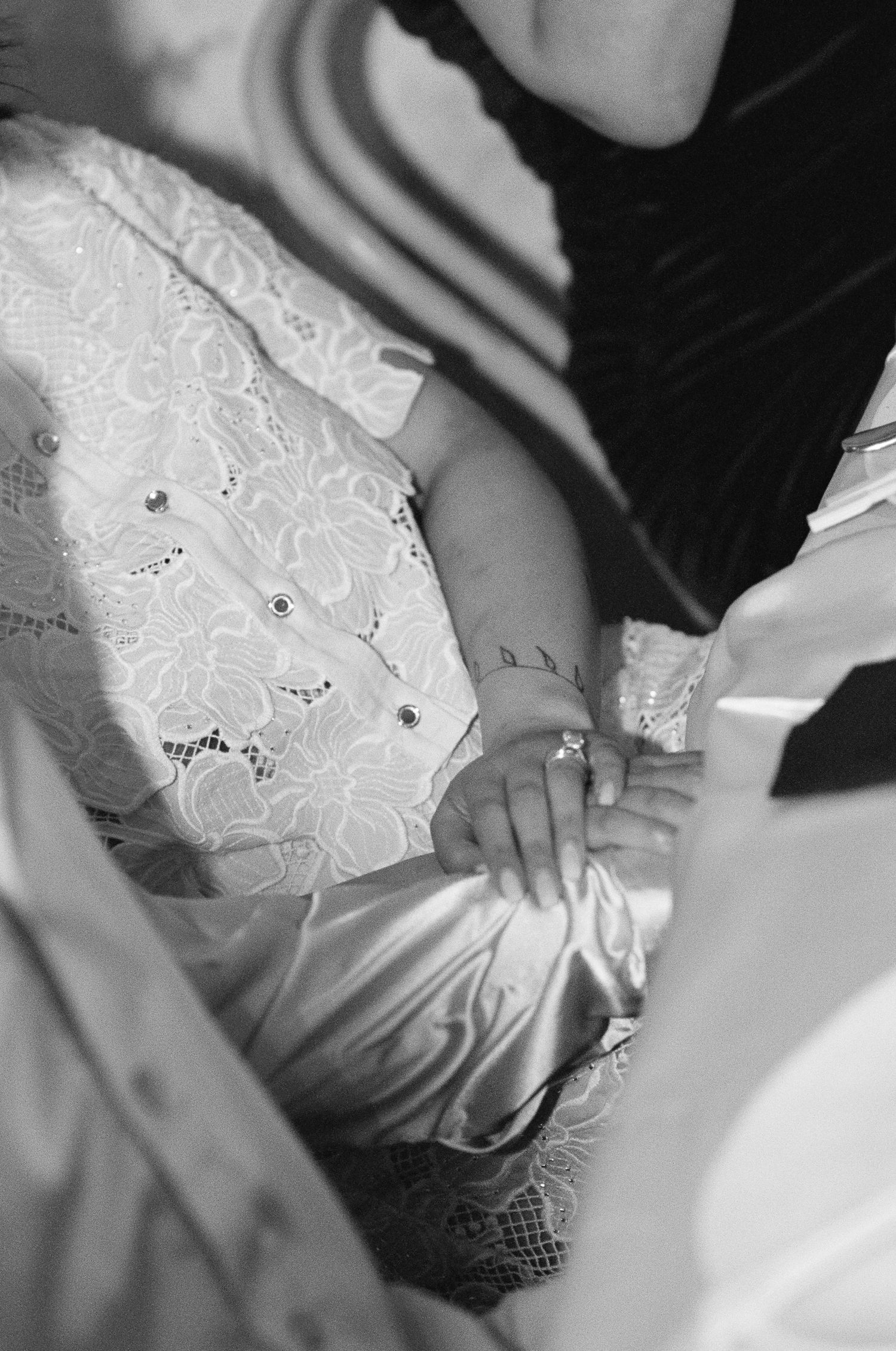 Black and white photo of a baby's small hand resting on a satin blanket, wearing a white lace outfit with jeweled buttons and a ring.