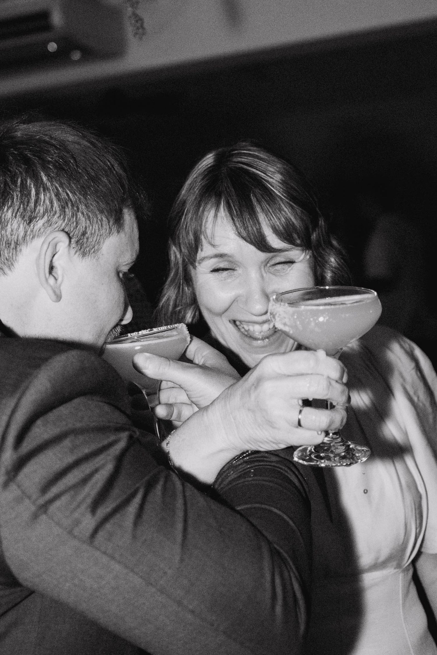 A joyful black and white photo of two people sharing cocktails and laughing together at a party.