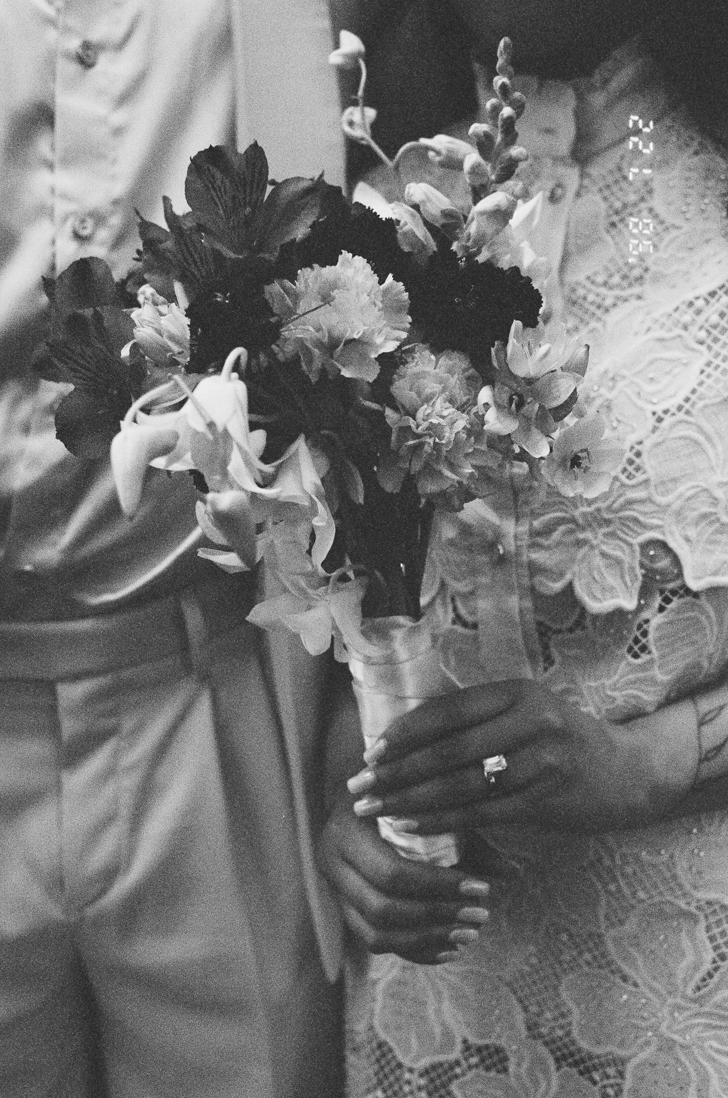 Black and white photo of a bride in a lace dress holding a wedding bouquet of lilies and mixed flowers beside a groom in light trousers.