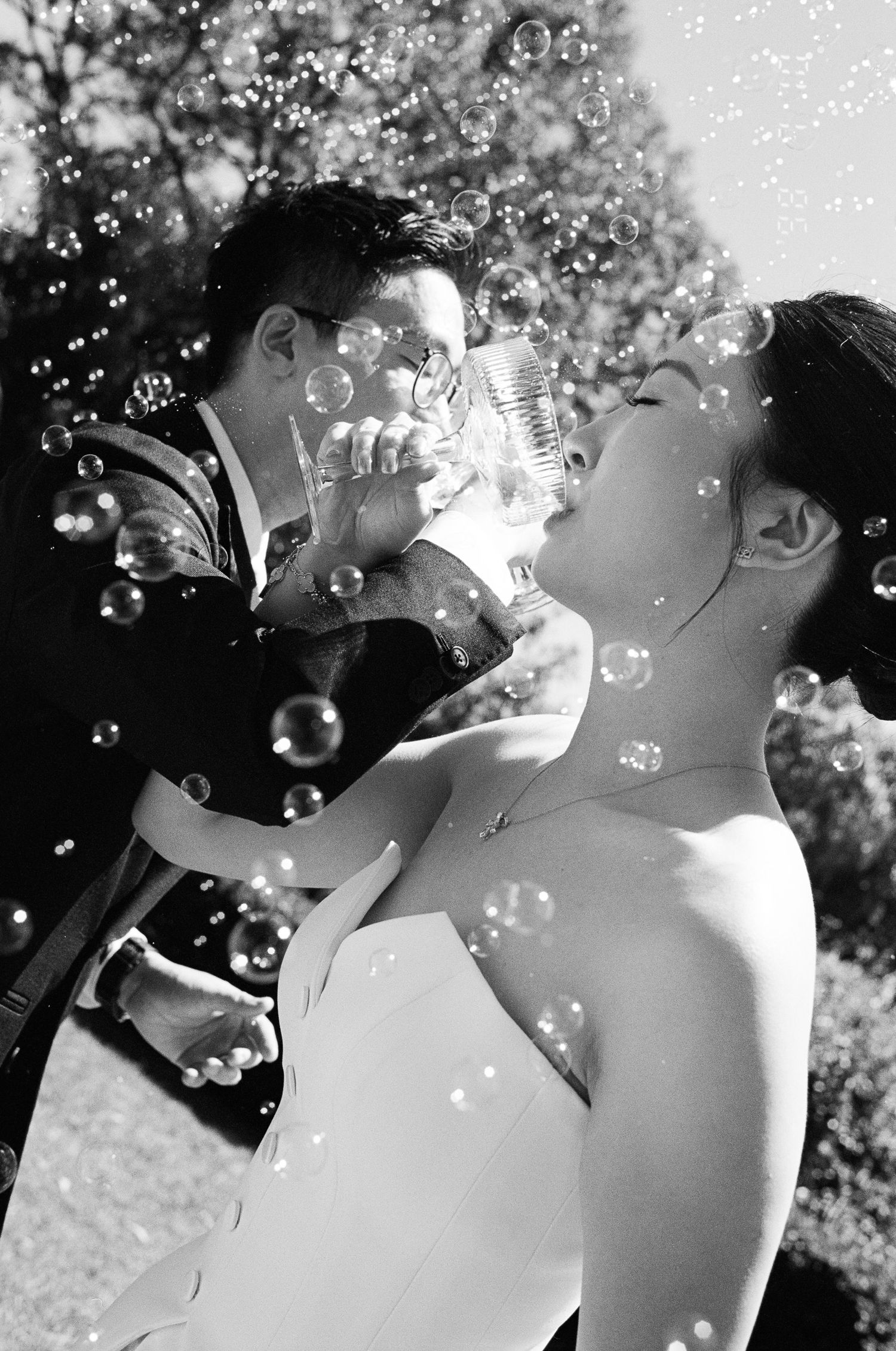 Black and white photo of a groom in a suit giving his bride in a strapless gown a drink while soap bubbles float around them.