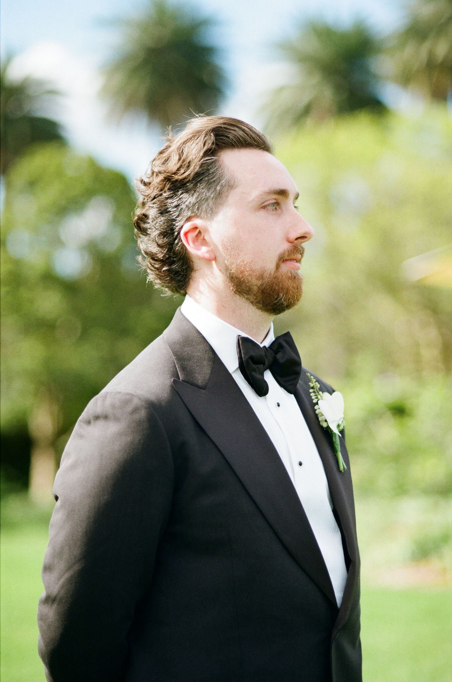 Groom in a black tuxedo and bow tie with a white boutonniere stands in profile in a sunny garden with palm trees.