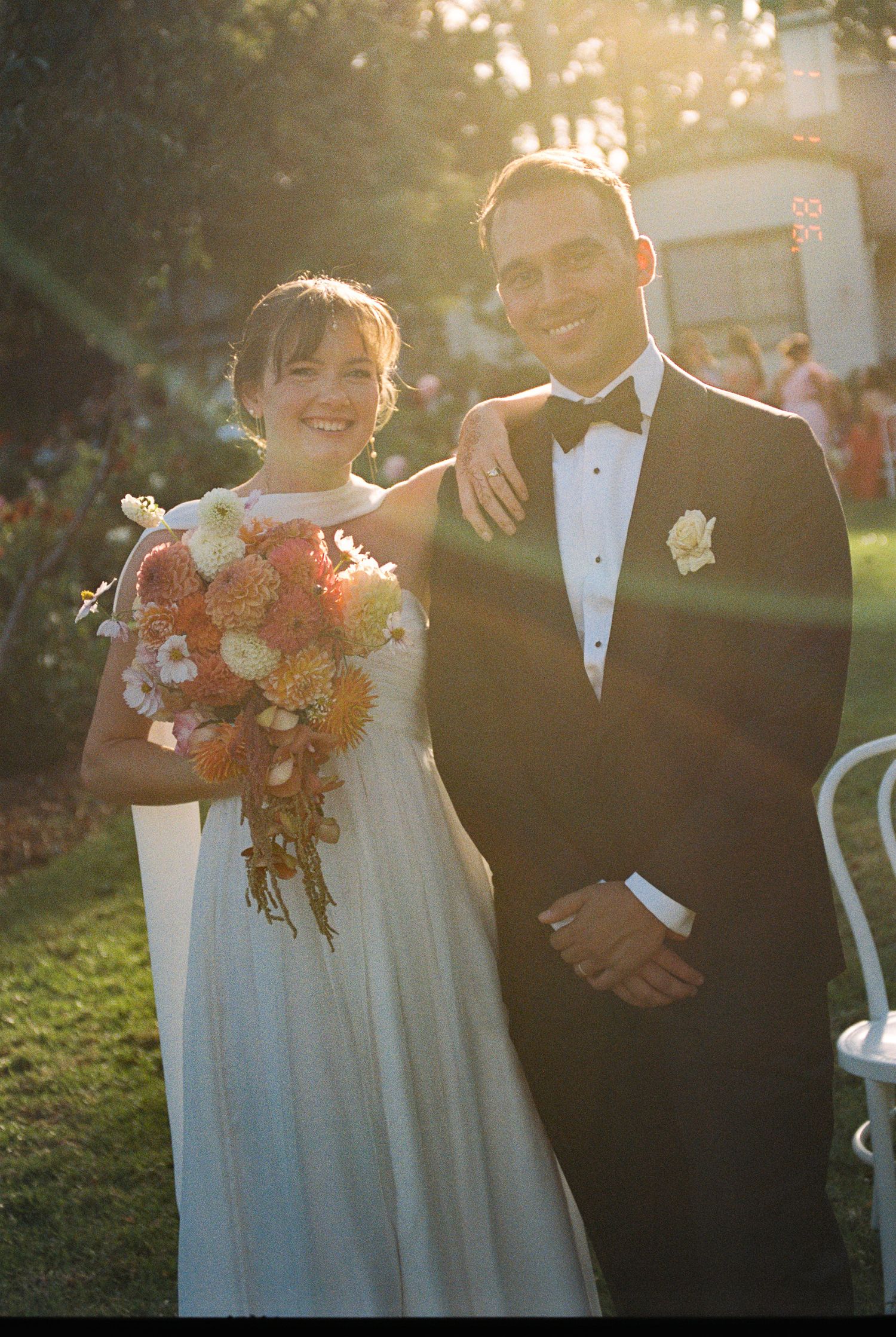 Smiling bride in a white gown holding an orange and peach dried flower bouquet beside a groom in a black tuxedo at a sunlit outdoor wedding.