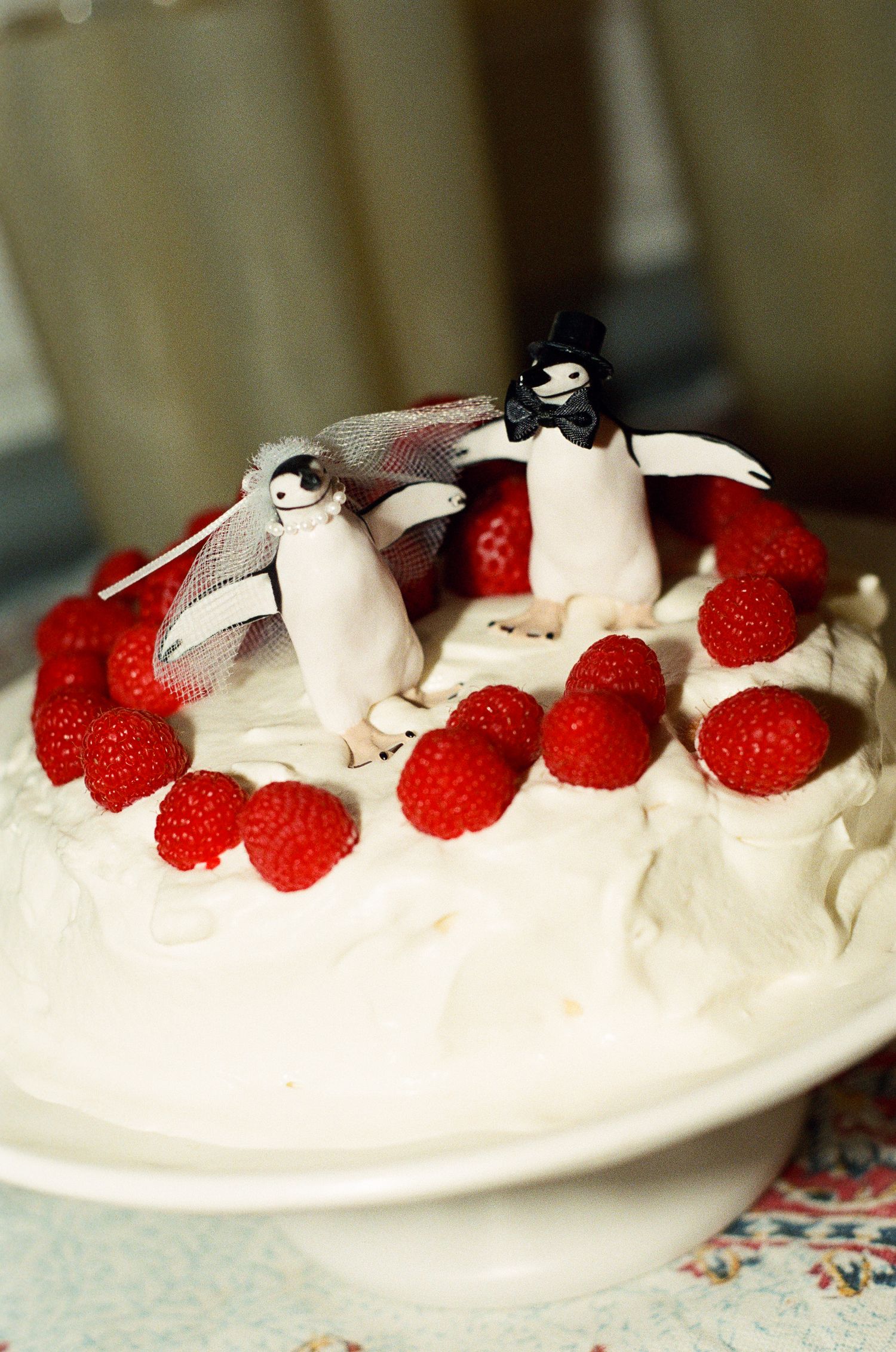 Two penguin figurines dressed as a bride with a veil and a groom with a top hat stand on a whipped cream cake with fresh raspberries.
