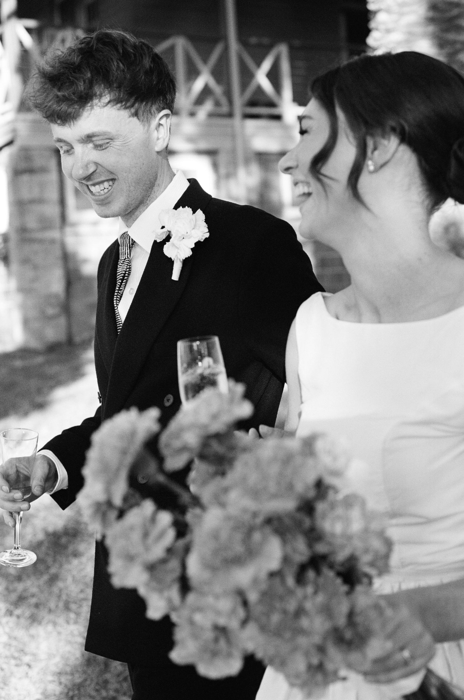 Black and white photo of laughing bride and groom holding champagne glasses and a floral bouquet.