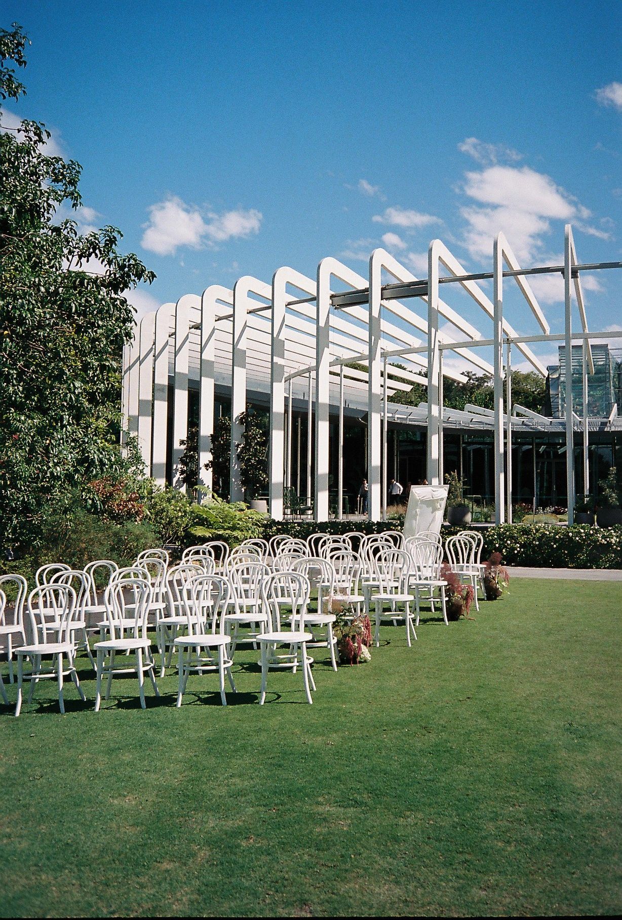White outdoor chairs arranged in rows on a green lawn facing a modern architectural structure with angled beams.
