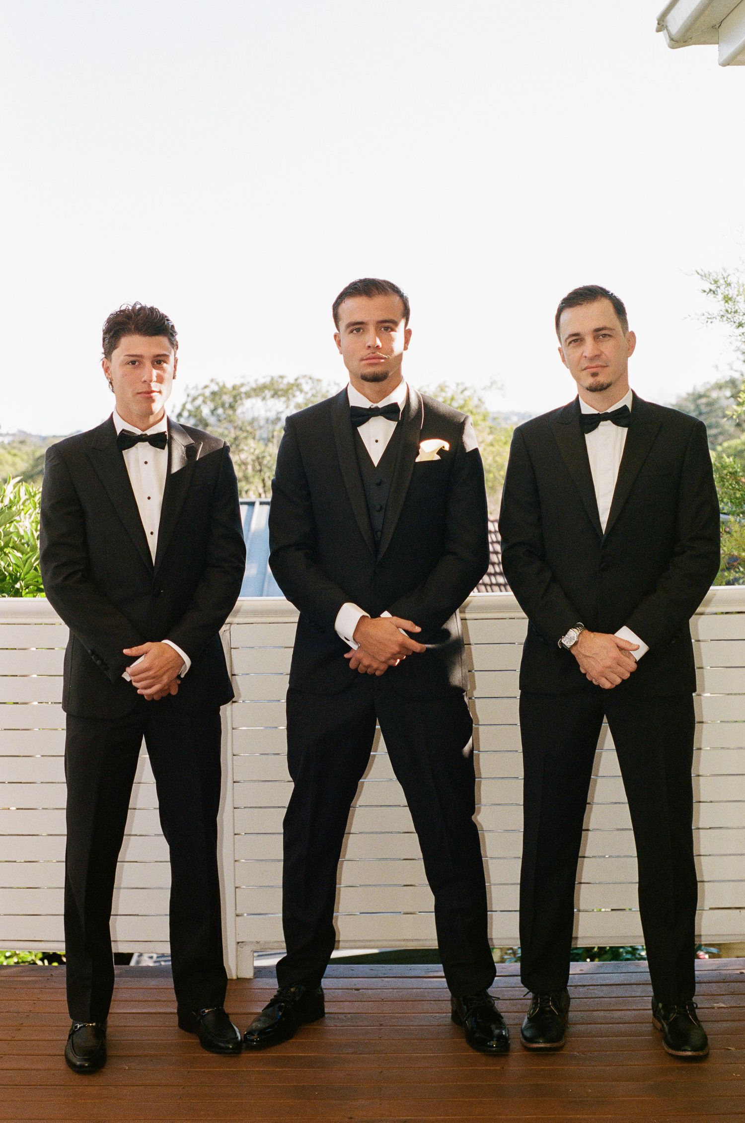 Three groomsmen in black tuxedos and bow ties stand side by side on a wooden deck with a white fence and trees behind them.